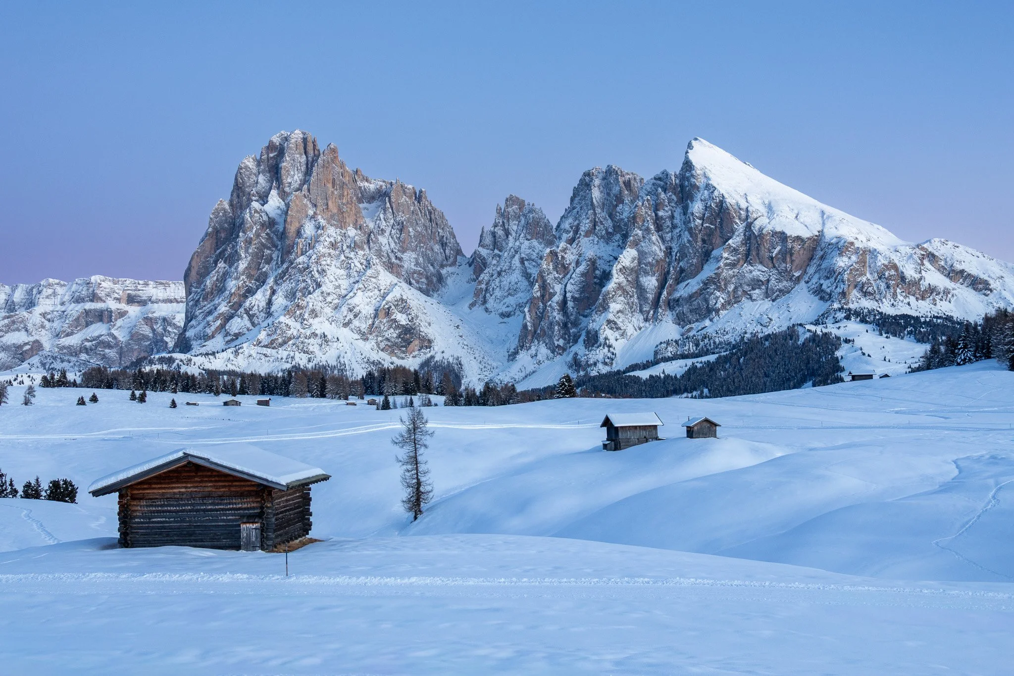 Snow-covered mountain landscape with several small wooden cabins scattered across the snow, and a clear blue sky overhead.