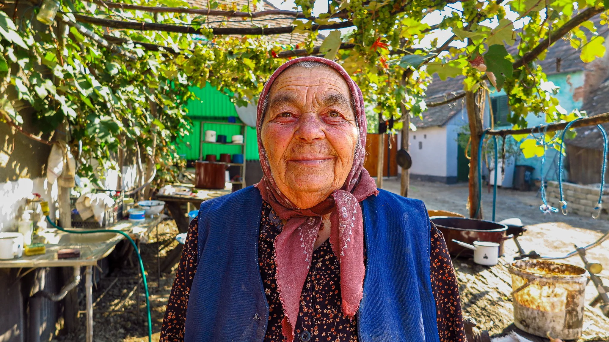 Smiling elderly woman wearing a headscarf and a blue vest standing outdoors in a rural yard with trees, a bicycle, and various household items in the background.