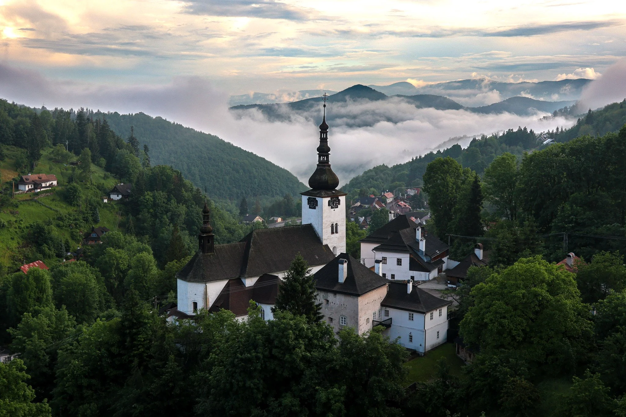 A church with a tall steeple in a mountainous landscape with fog, green trees, and a small village.