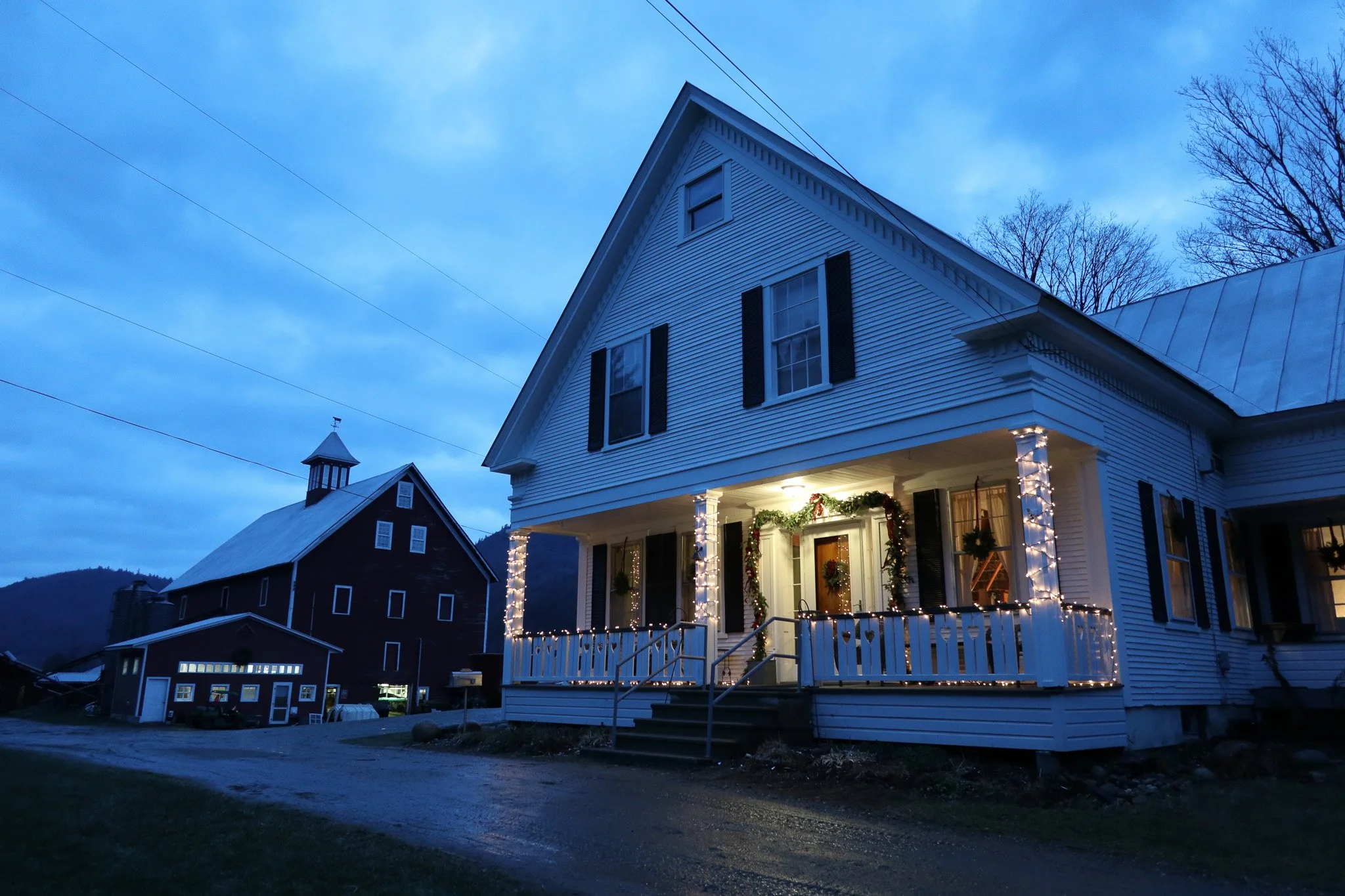 A white house decorated with Christmas lights and wreaths on the front porch, at dusk with a dark blue sky and a nearby barn in the background.