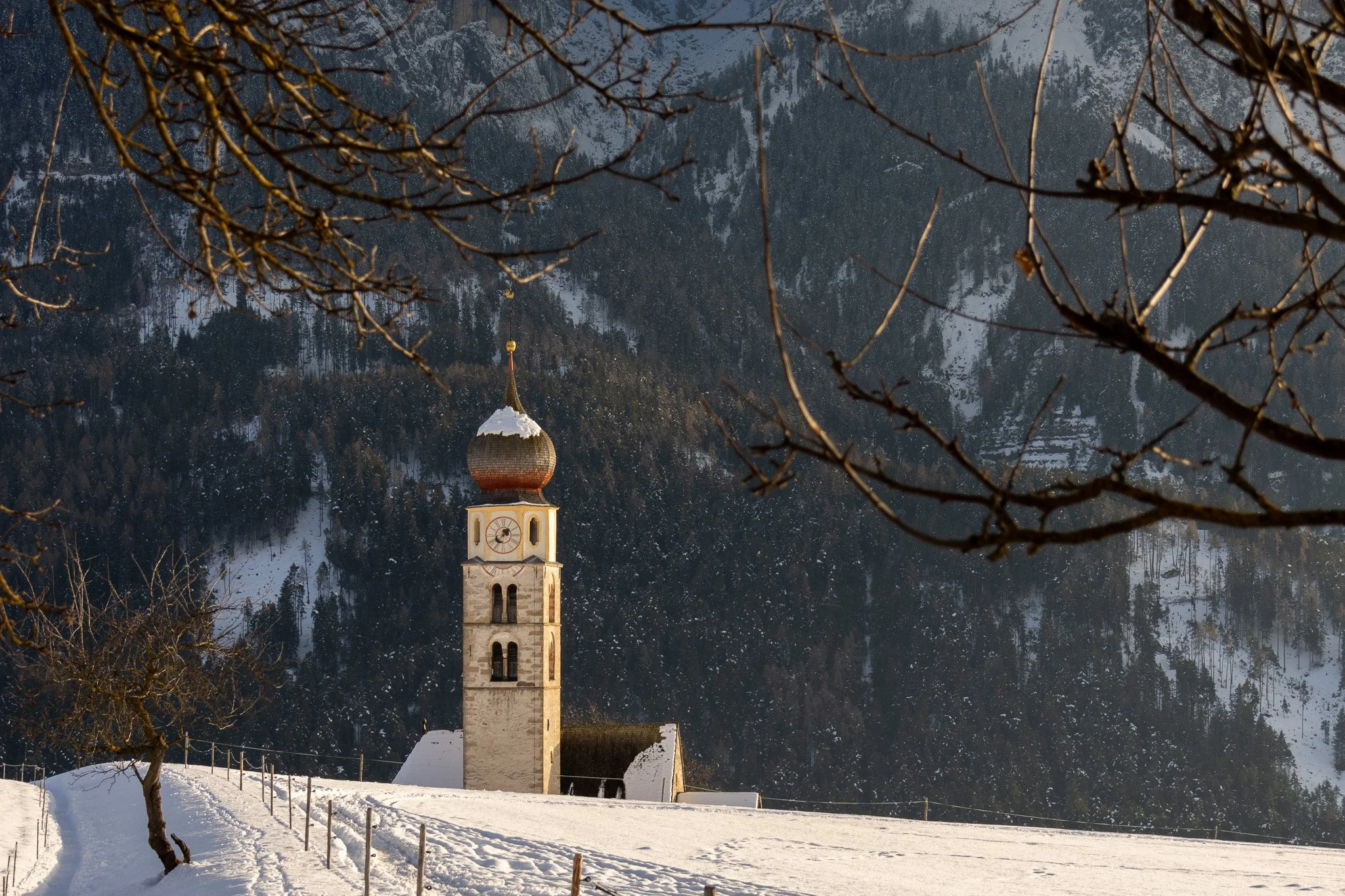 A snow-covered landscape with a church tower featuring a clock, set against a backdrop of snow-covered mountains and trees, with some bare tree branches framing the scene.