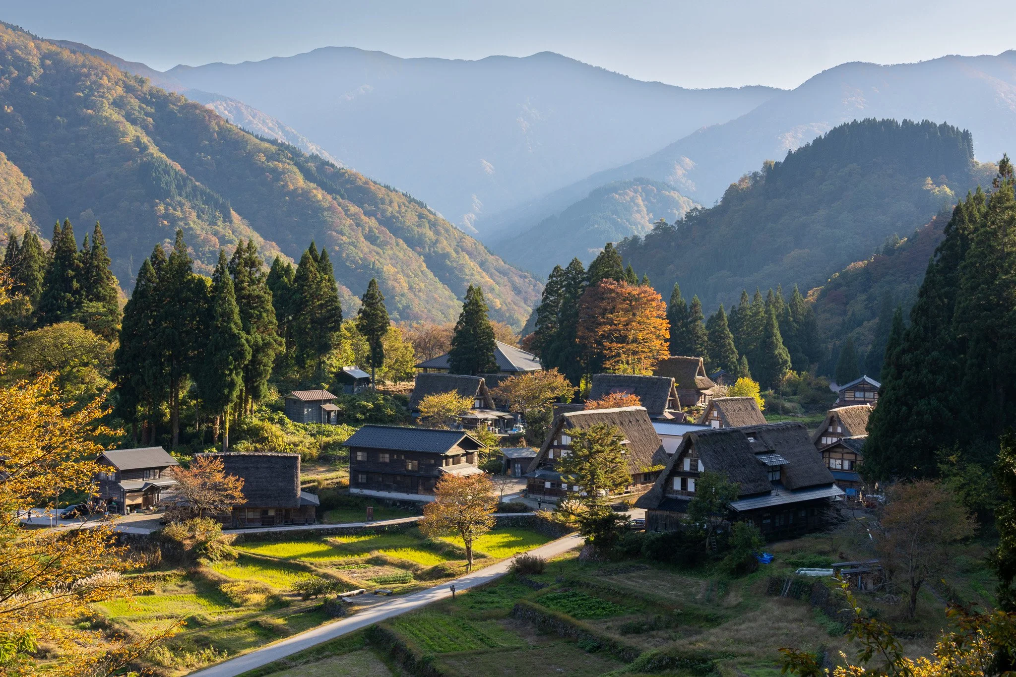 Scenic view of a traditional Japanese village nestled in a mountainous region with lush green forests and autumn foliage, under a clear sky.