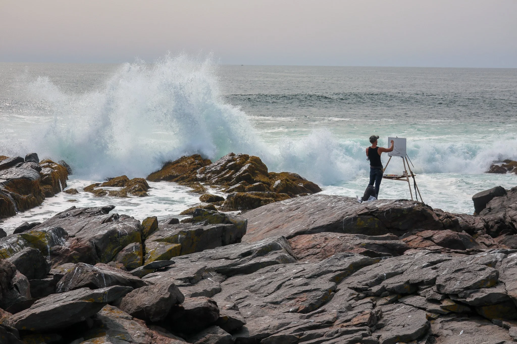A person wearing a black sleeveless top and jeans, standing on rocks by the ocean, painting on an easel as waves crash nearby.