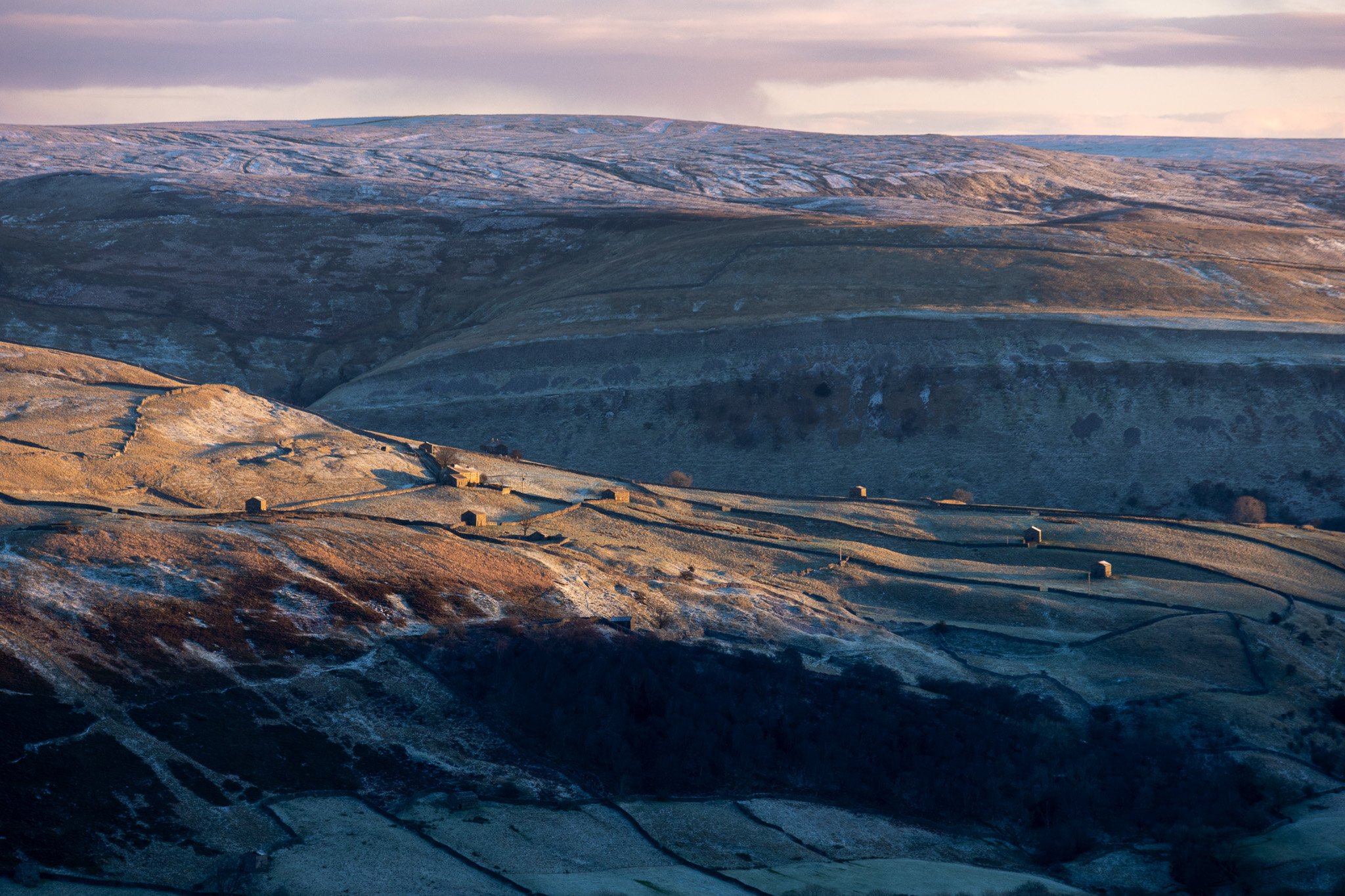 Hilly landscape with patches of snow, small houses, and winding roads at sunset or sunrise.