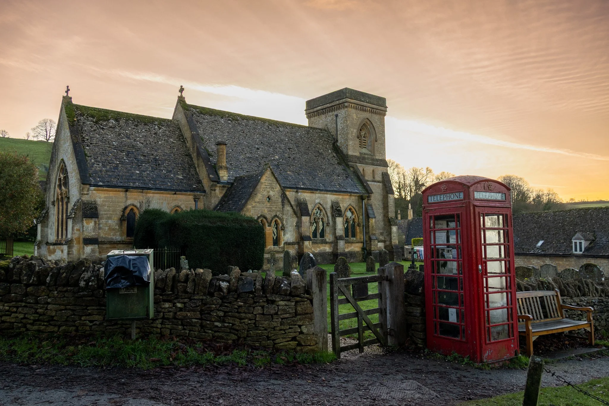 A stone church with a grey roof and a red telephone booth outside, surrounded by a low stone wall and a park bench, with a sunset sky in the background.
