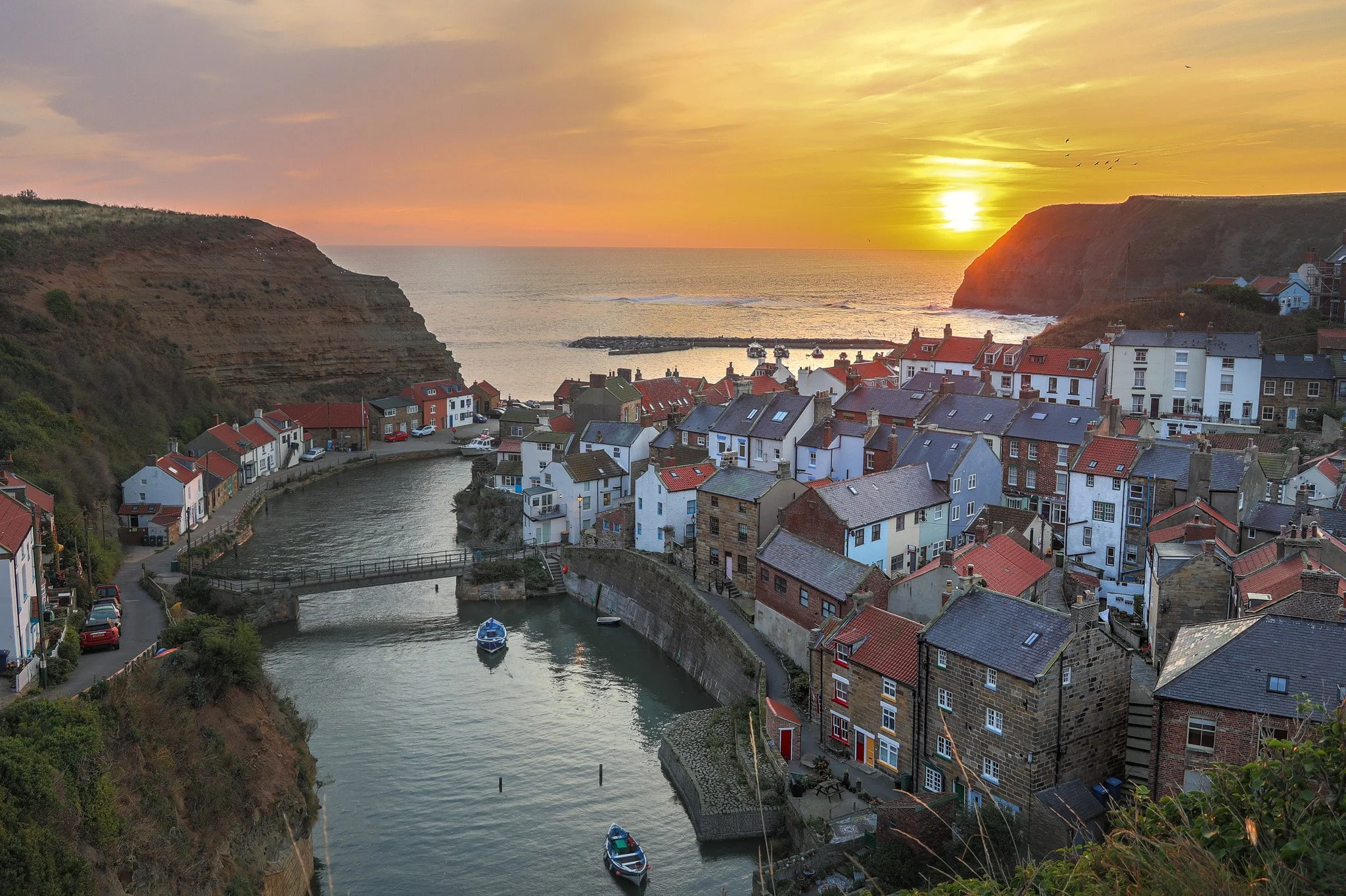 A coastal village at sunset with colorful houses lining a narrow harbor, surrounded by cliffs, and a calm sea in the background.