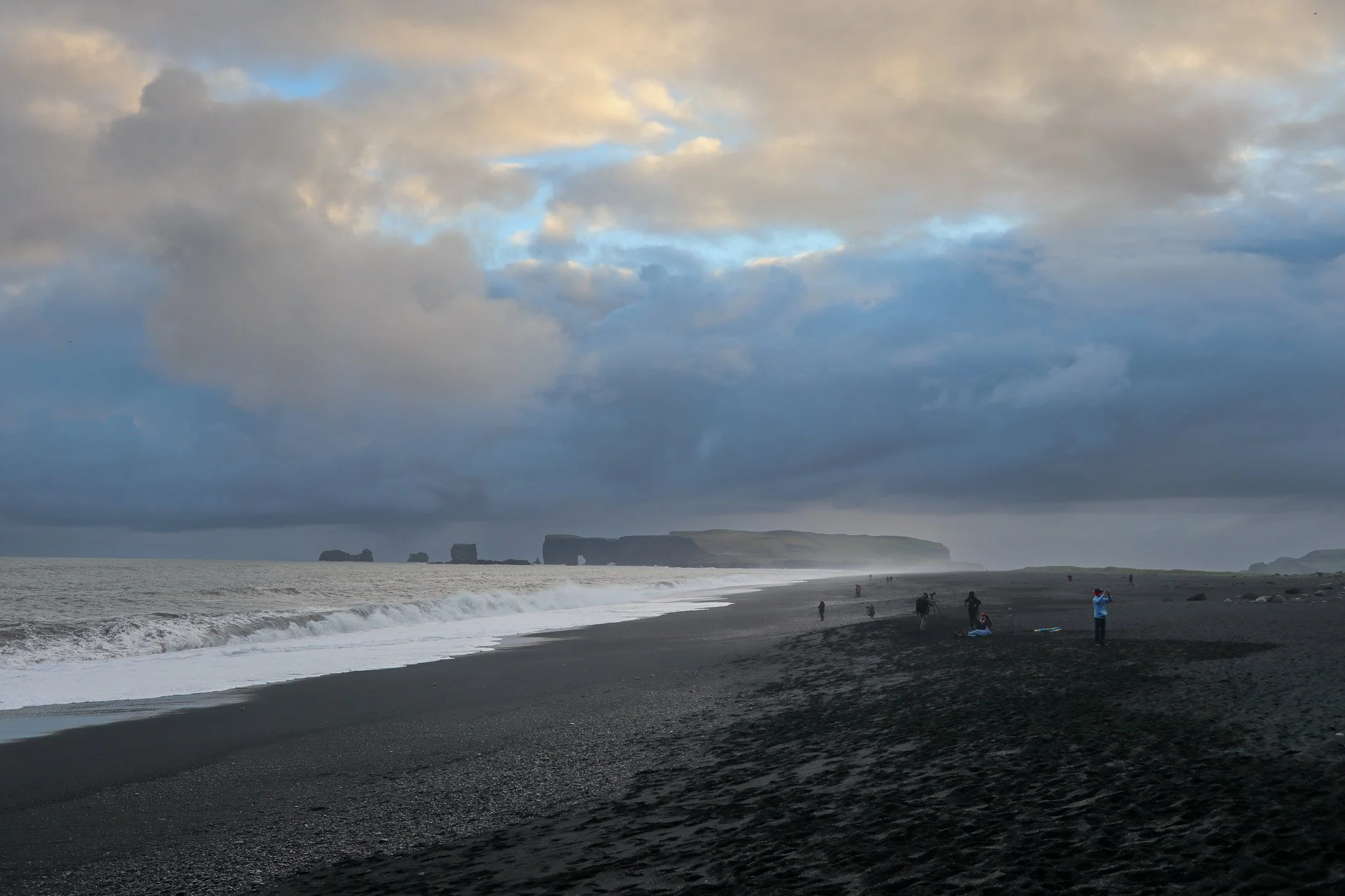 A black sand beach with people walking and taking photos, ocean waves, rocky cliffs in the distance, and a cloudy sky.