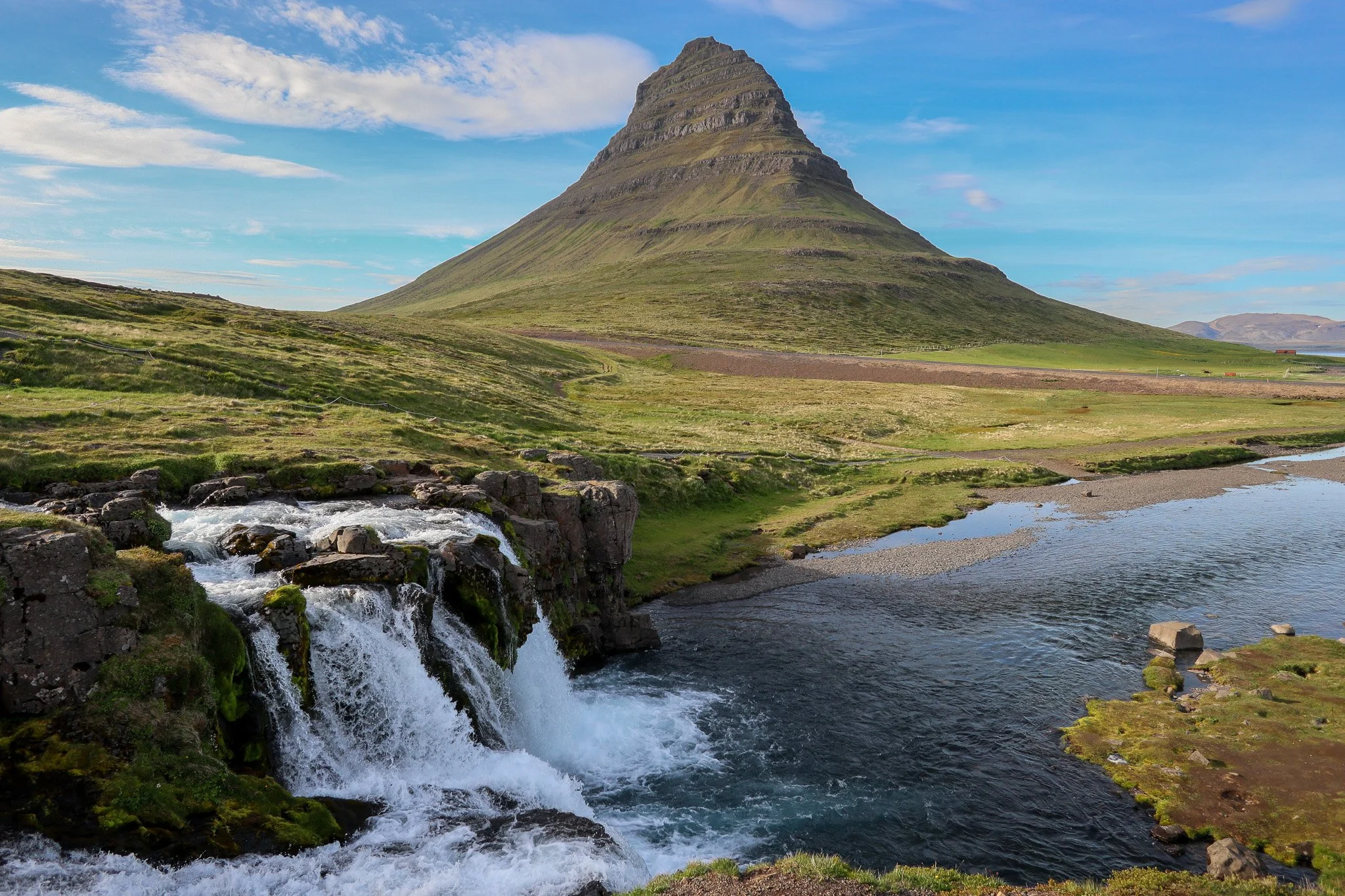 A scenic landscape featuring a tall, conical mountain with grassy slopes, a small waterfall flowing into a river in the foreground, and a partly cloudy sky.