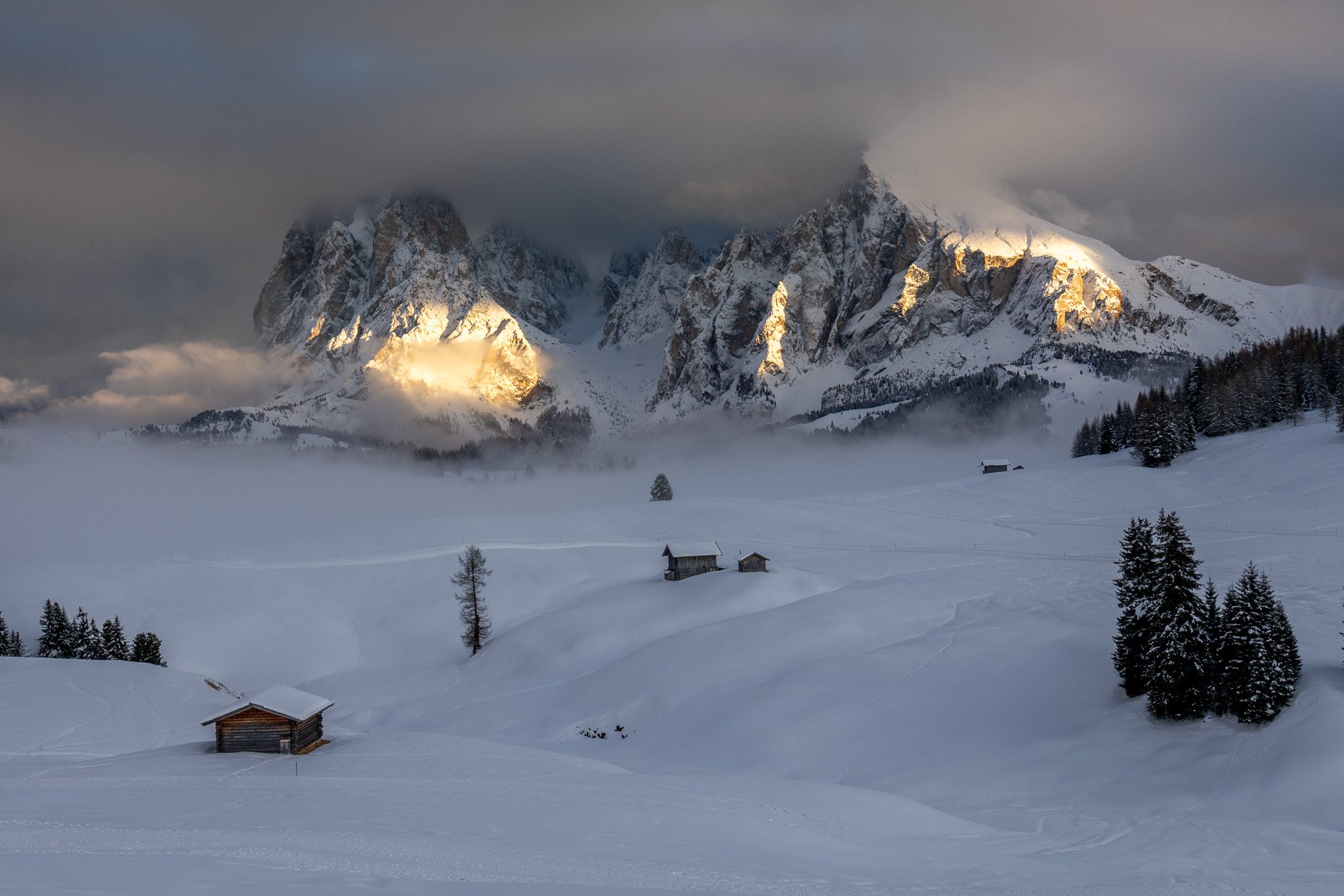 Snow-covered landscape with a few small wooden houses, snow-covered trees, and mountain peaks illuminated by sunlight in the background under cloudy sky.