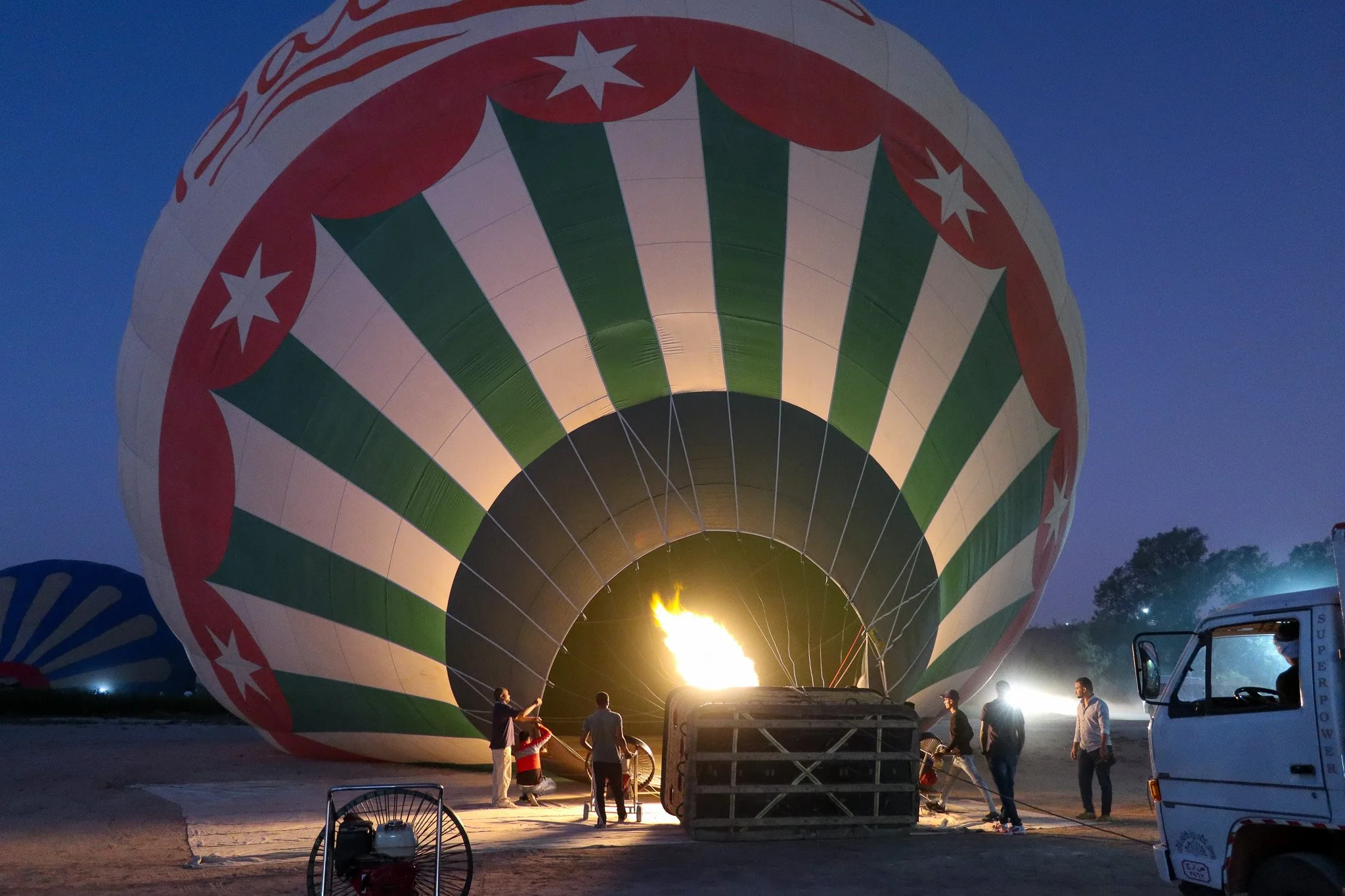 People preparing a hot air balloon for flight at dusk, with the flame heating the balloon's interior and a propane tank nearby.