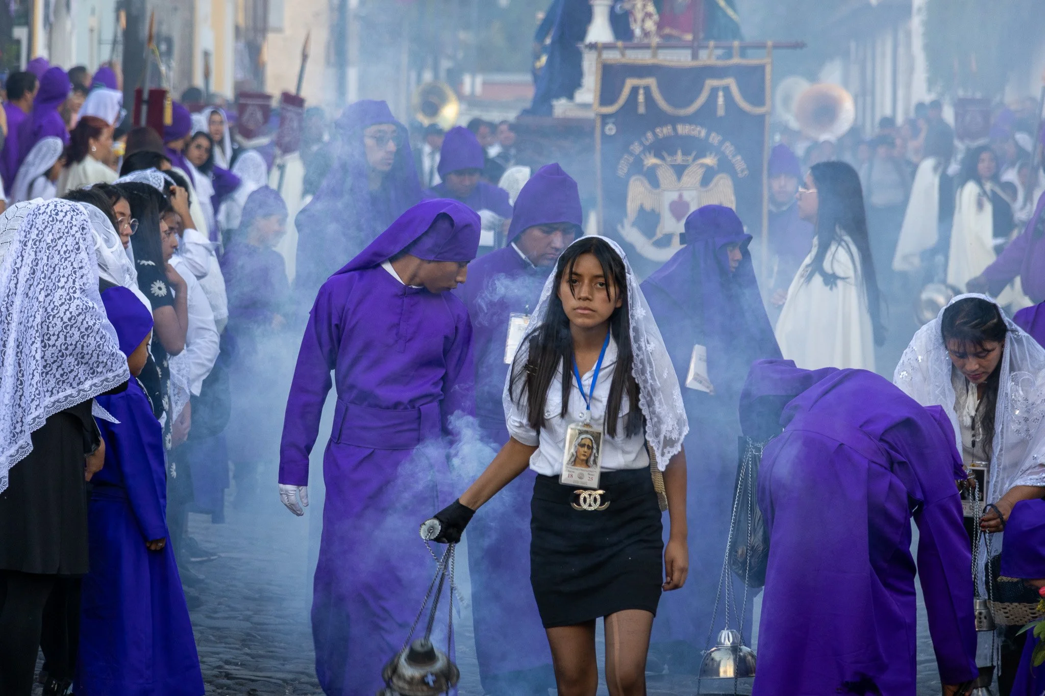 People participating in a religious procession or ceremony, many dressed in purple and white, with some women wearing mantillas, and holding lanterns or incense holders, on a cobblestone street.