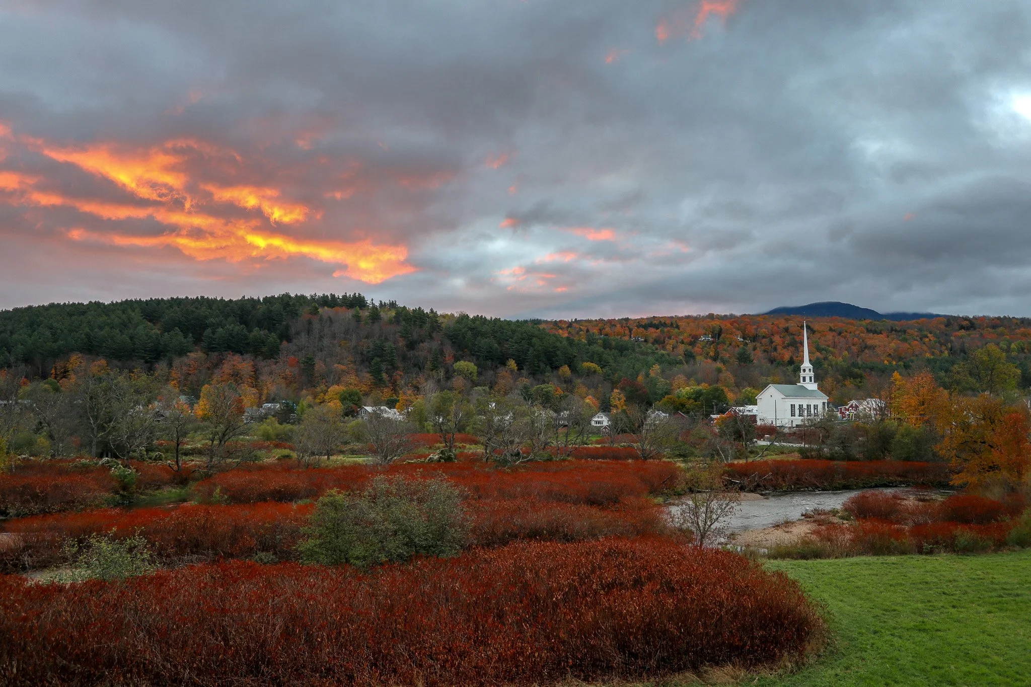 A scenic landscape with colorful autumn trees, a river, a white church with a steeple, and a cloudy sky with hints of orange sunset.