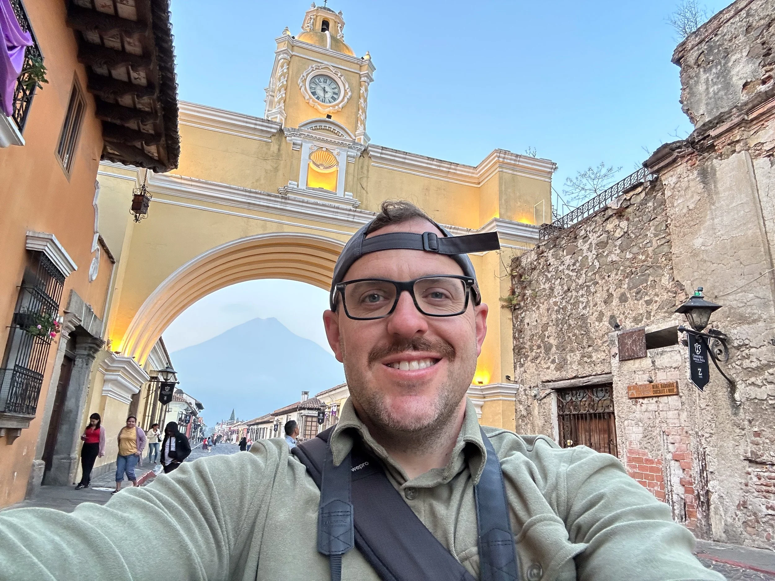 Smiling man wearing glasses, a backward baseball cap, and a backpack taking a selfie in a historic city with colorful buildings, a large clock tower, and a volcano in the background.