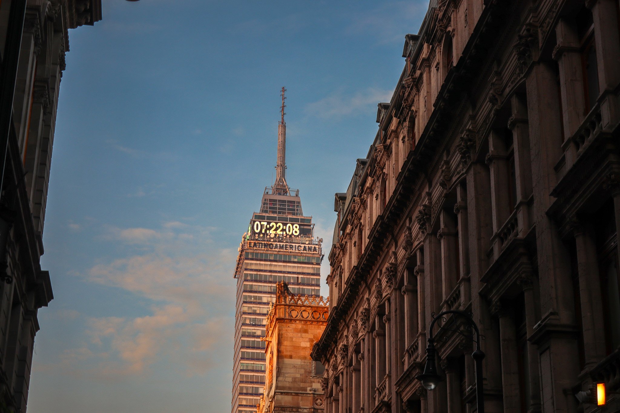 City street view with historic buildings on both sides, and the Torre Latinoamericana skyscraper in Mexico City visible in the background with a digital clock display showing 07:22:08 at the top of the building.