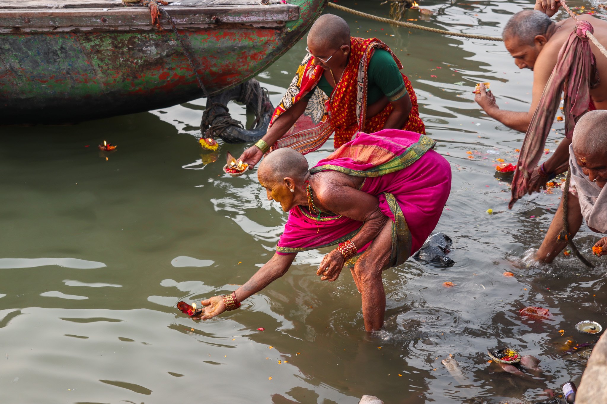 Women and men performing a water ritual, standing in a river near a boat, offering and releasing flower petals and small ritual items.