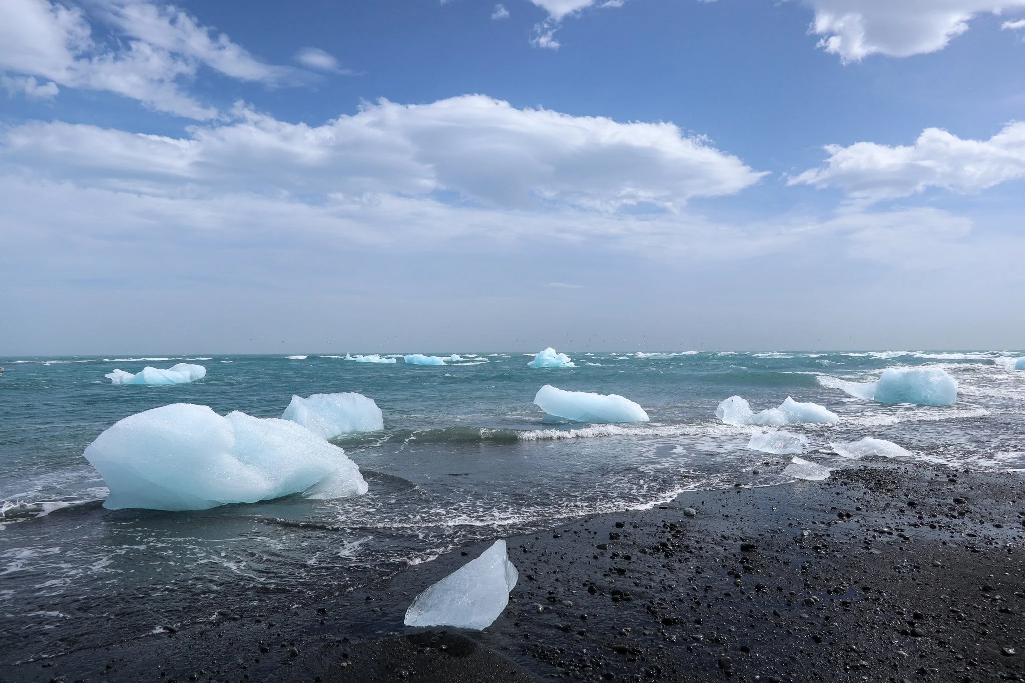 Ice chunks scattered on a black pebble beach with the ocean and cloudy sky in the background.