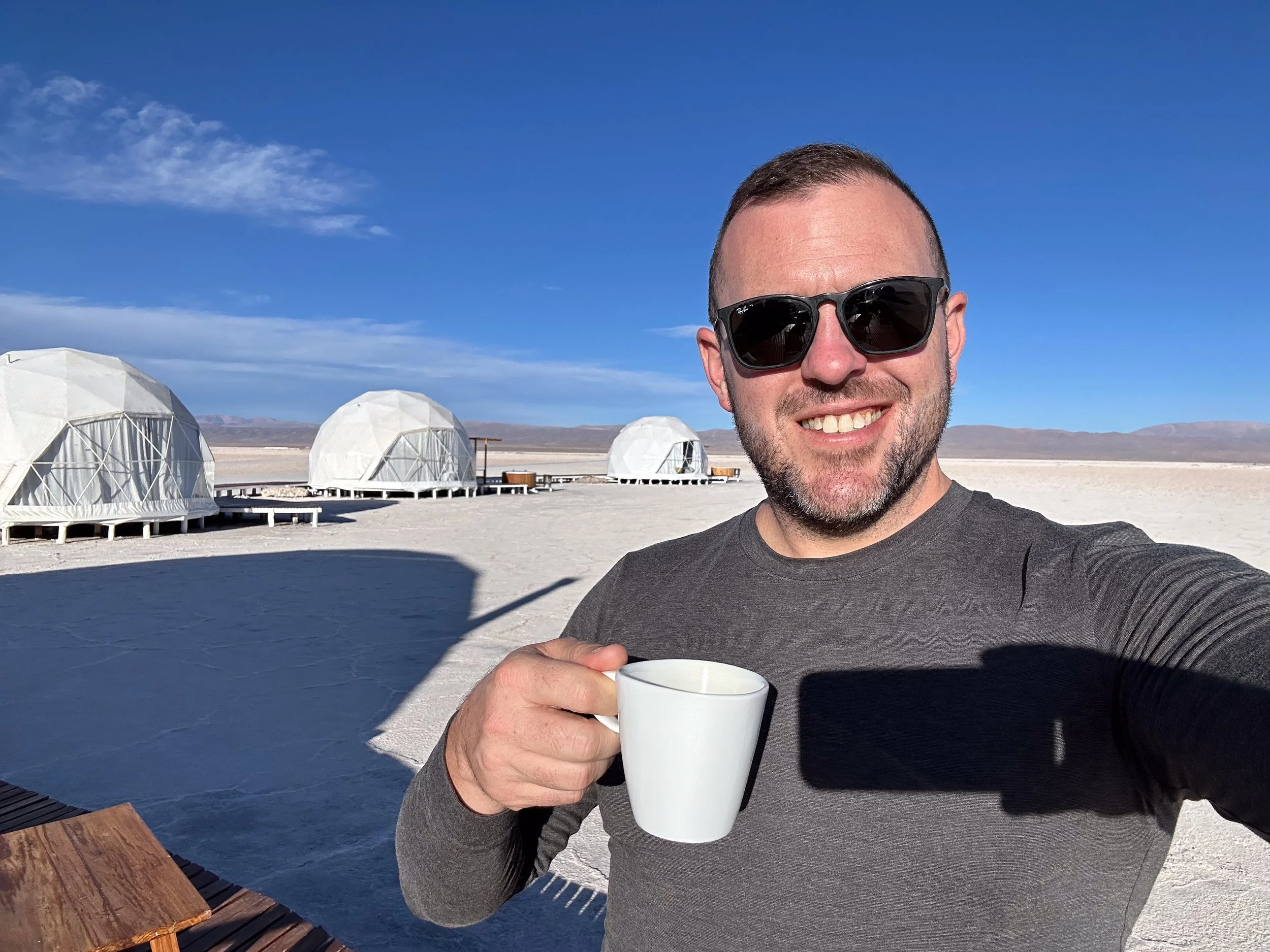 A man wearing sunglasses and a gray long-sleeve shirt smiling while holding a white mug outside on a sunny day with geodesic dome structures in the background on a desert landscape.