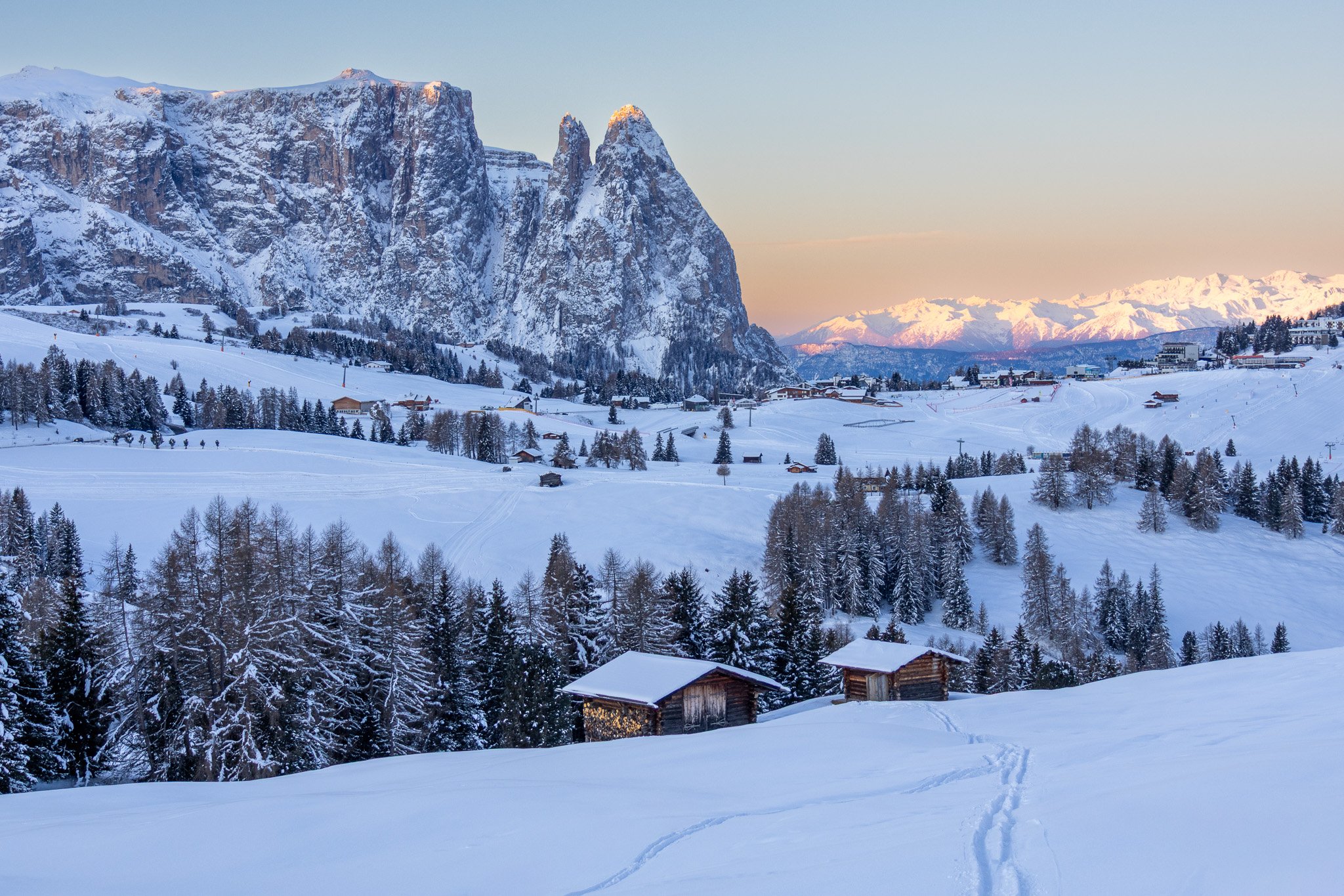 Snow-covered mountains and a valley with small wooden houses, trees, and ski trails during sunset, with a clear sky and distant snow-capped peaks.