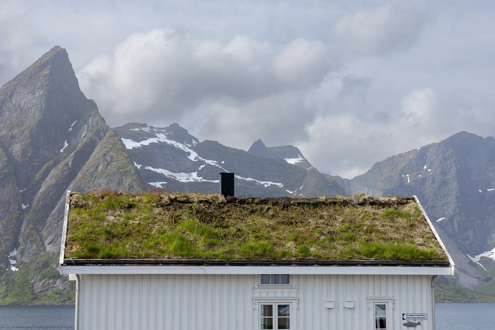 A house with a green roof and white exterior, situated near a body of water, with mountains in the background and a cloudy sky.