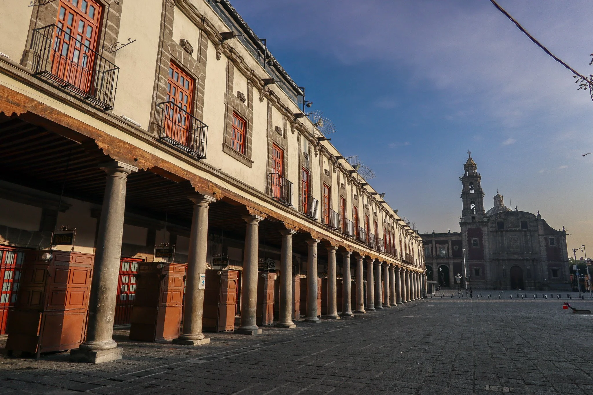 Empty historic square with a large multi-story building supported by columns and a church with a bell tower in the background, under a partly cloudy sky.