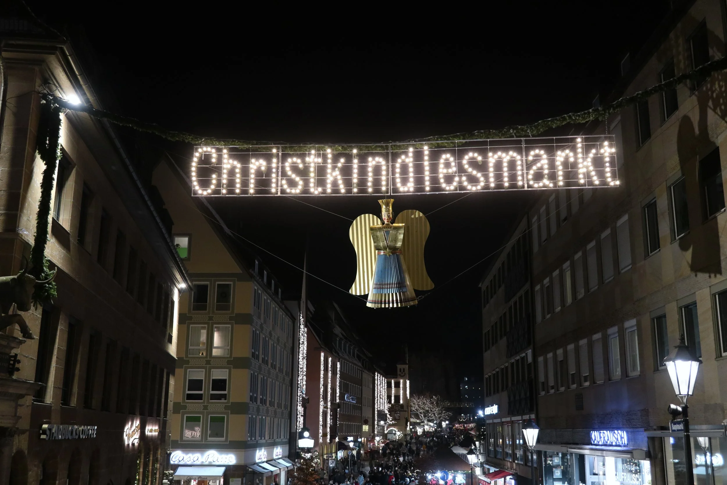 Night view of a street decorated with Christmas lights, including a high-lit sign displaying 'Christmasmarket' and a large angel figure hanging below it.