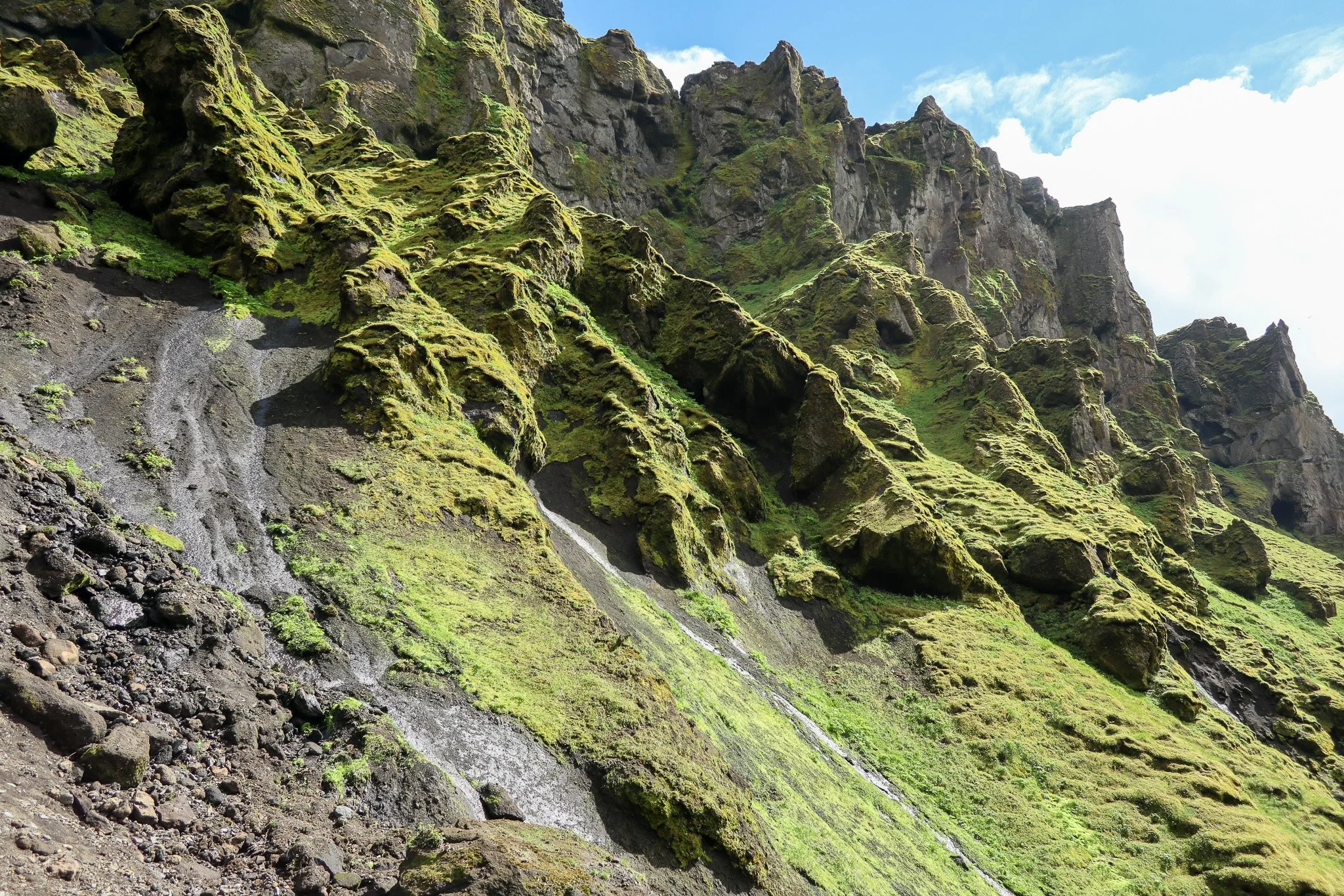A steep mountain hillside covered in green moss and grass with exposed dark rocks and a small waterfall.