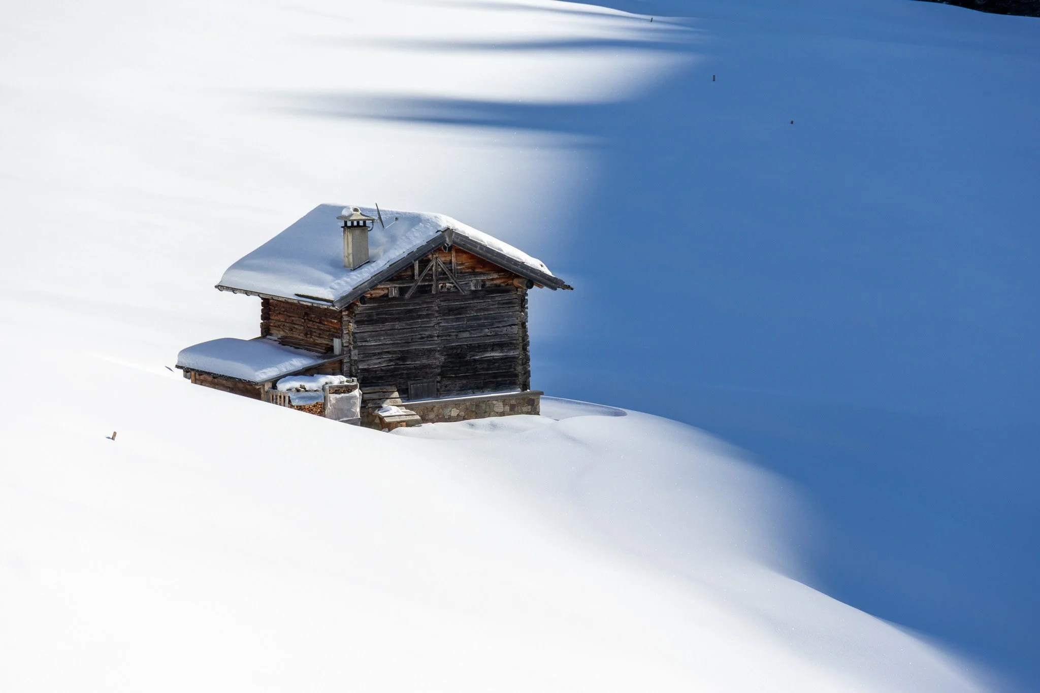 A lone wooden cabin covered in snow on a snowy landscape with a clear blue sky.