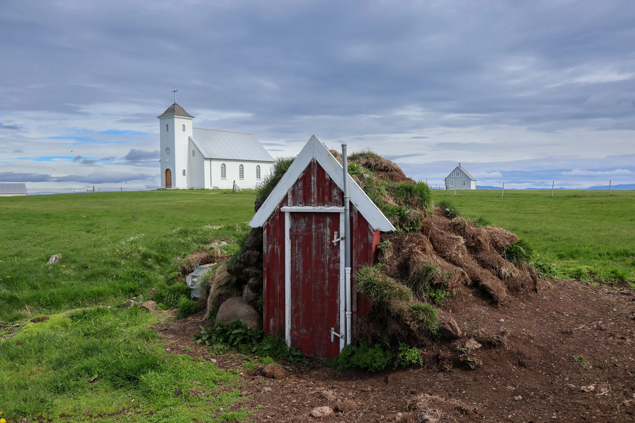 A small red and white wooden hut built into a grassy hill with a church and other buildings in the distance on a cloudy day.