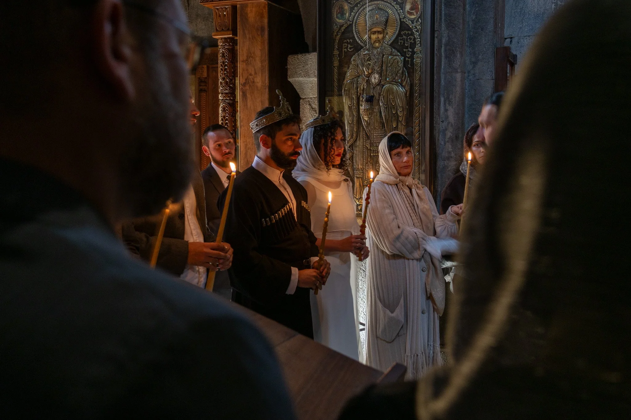 People participating in a religious ceremony inside a church, holding candles, with an icon of a saint on the wall behind them.