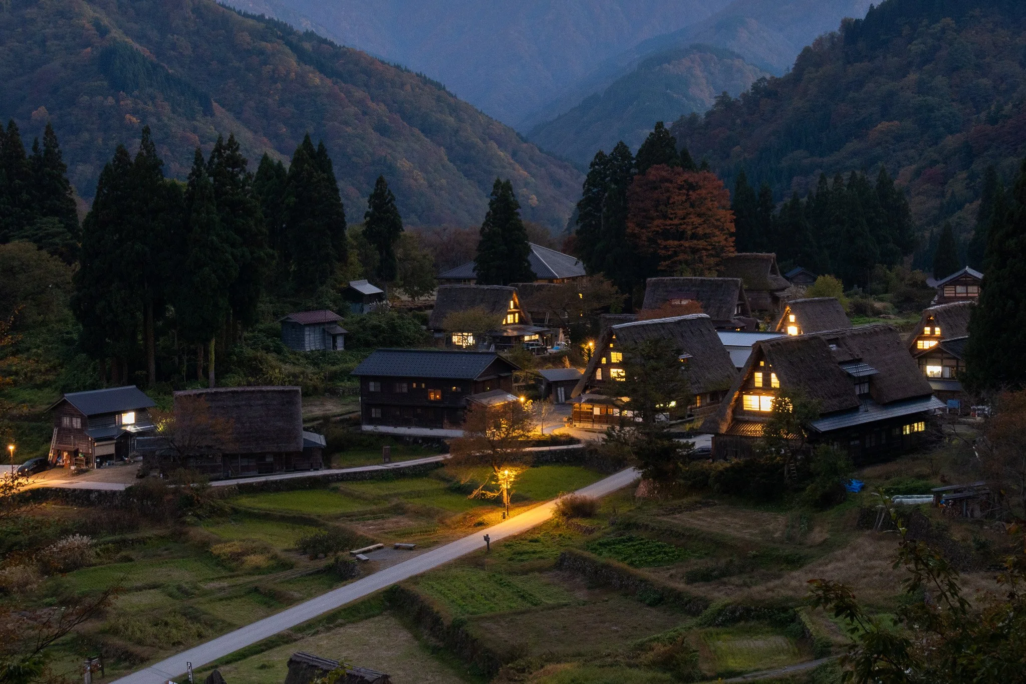 A scenic view of a tranquil village with traditional thatched-roof houses illuminated from within, nestled among lush green mountains and tall trees at dusk.