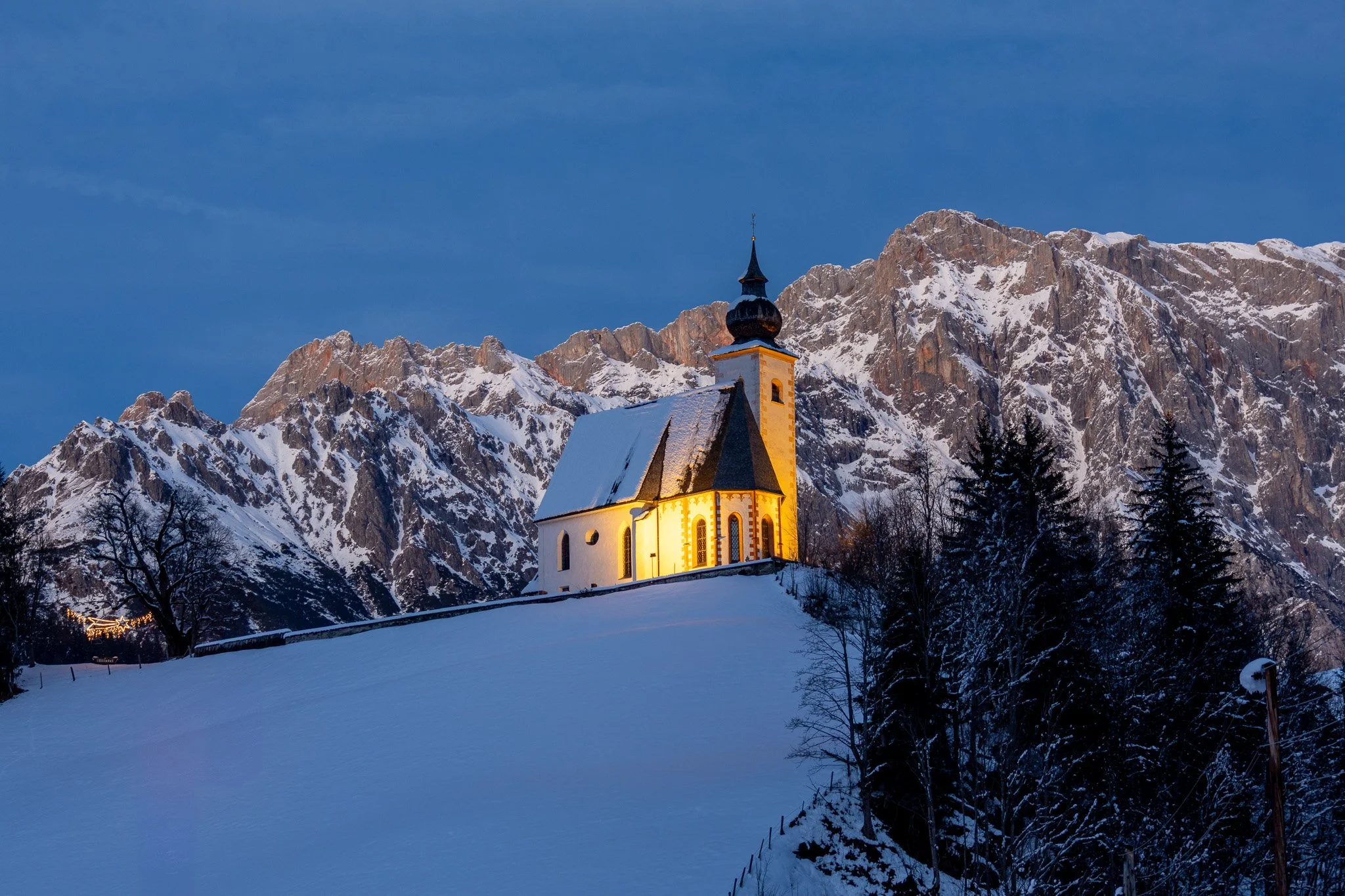 A small illuminated white church on a snowy hill with a mountain range in the background during twilight.