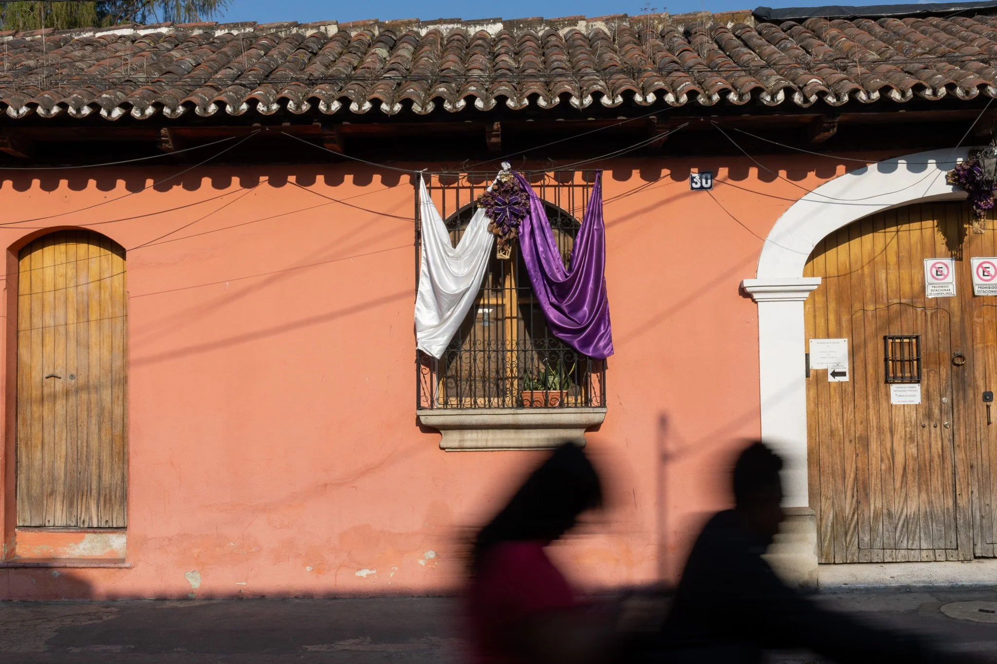 A peach-colored building with wooden doors and a small balcony with purple and white drapes hanging from the railing. The building has a tiled roof and a sign with the number 30. Two blurred people are walking in front of the building.