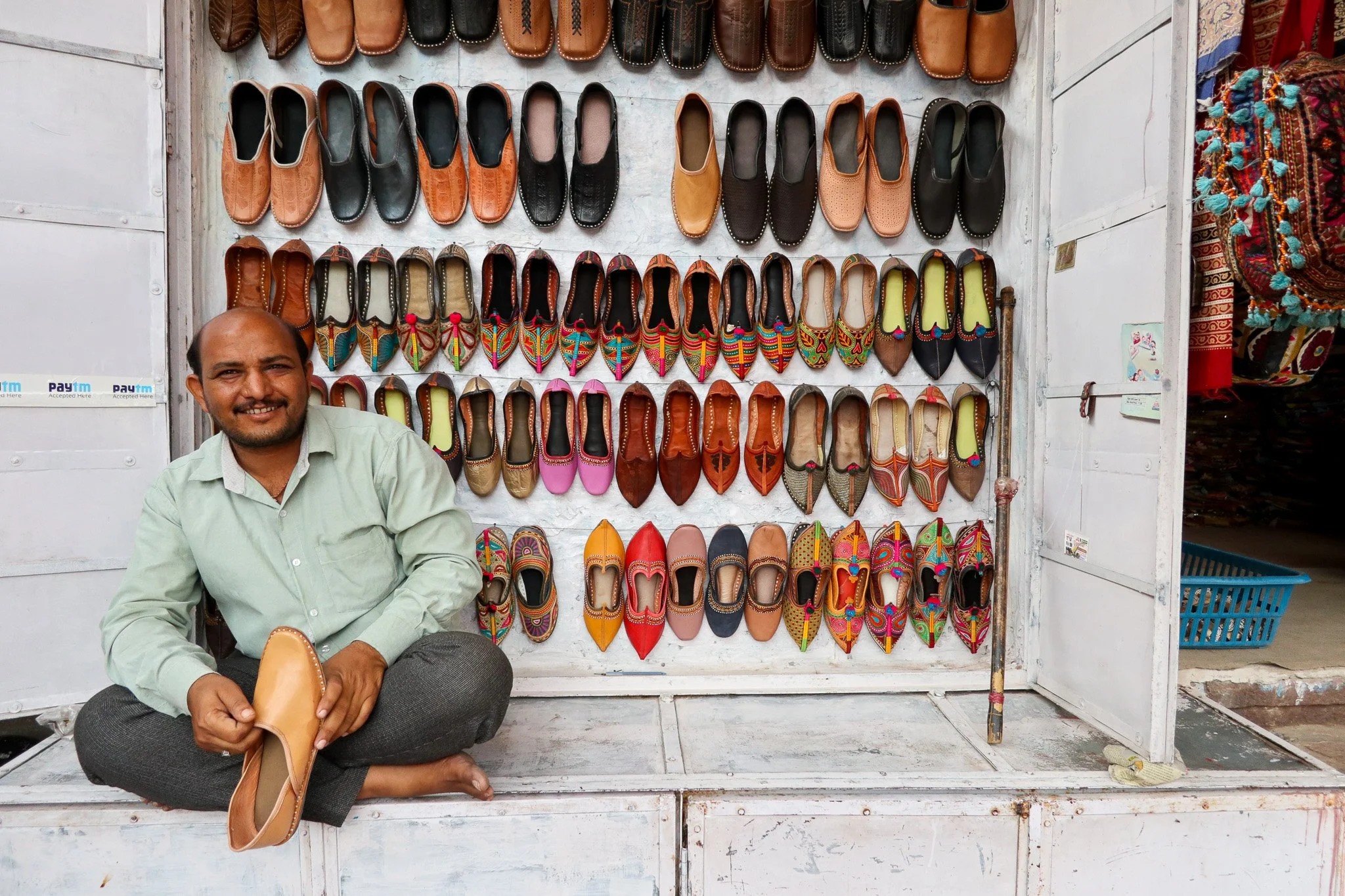 A man sitting cross-legged on a white wooden platform in front of a display of various colorful shoes at a street stall.