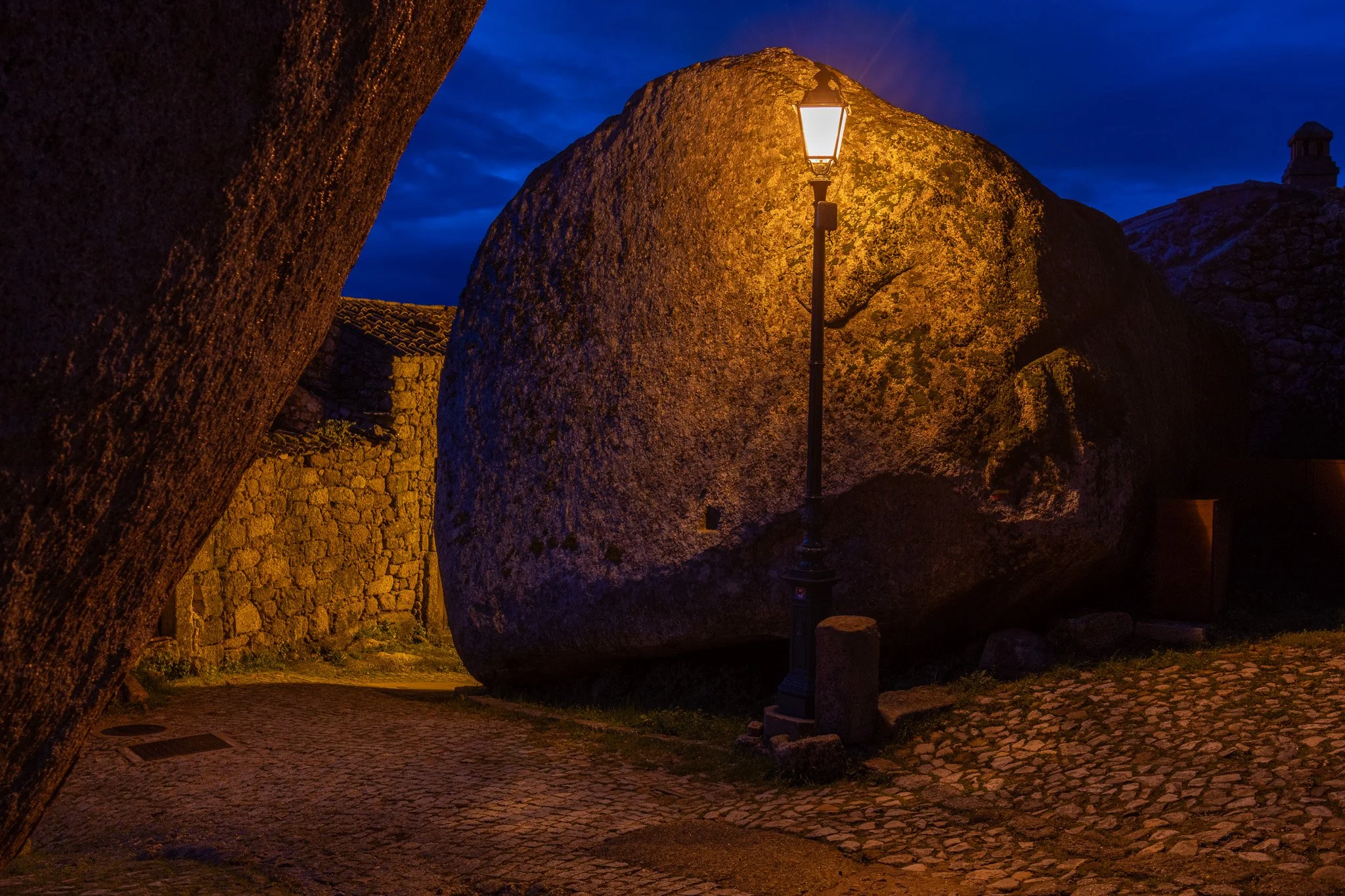 A large boulder rocks in a quaint village at night, softly illuminated by a streetlamp, with cobblestone streets and stone buildings around, under a dark blue sky.