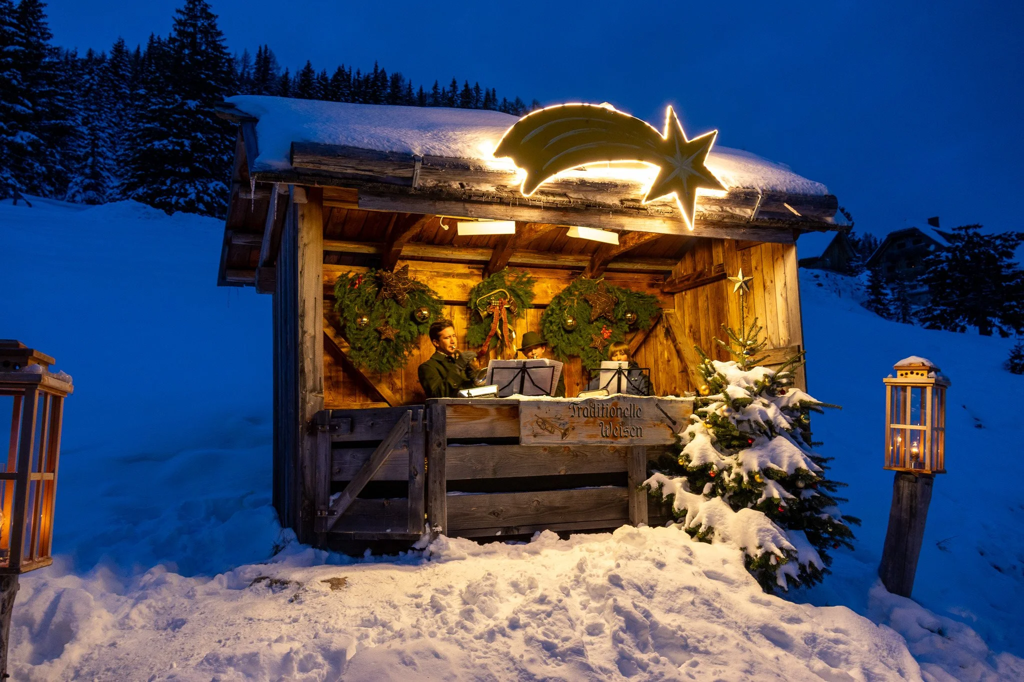 A nighttime outdoor scene of a wooden stage decorated with Christmas wreaths and plants, featuring three musicians playing wind instruments, with a snow-covered ground and Christmas tree, and lanterns illuminating the area.