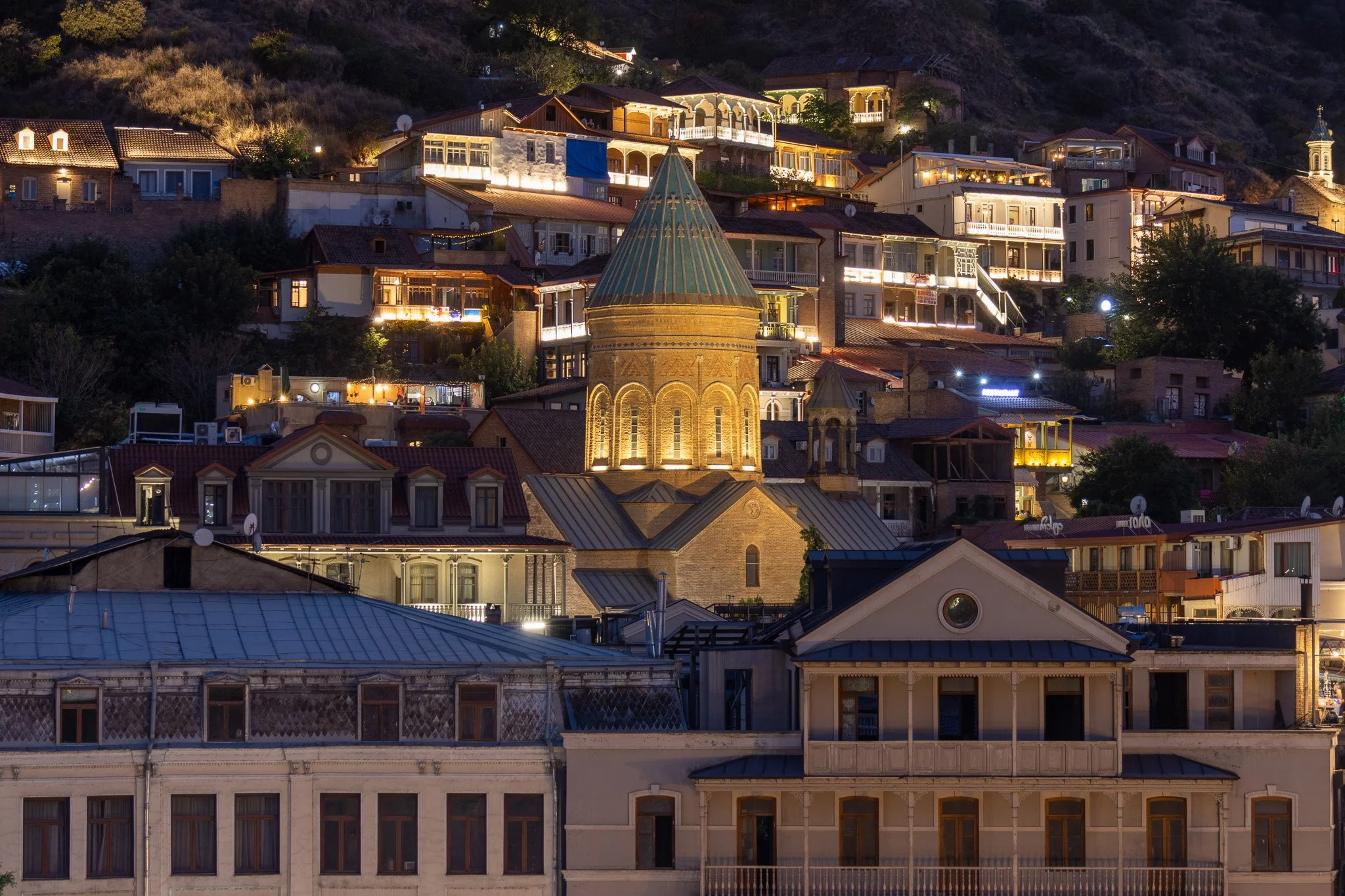 Nighttime view of a hillside city with various illuminated houses and buildings, featuring a prominent historic brick church with a tall, pointed green roof in the center.