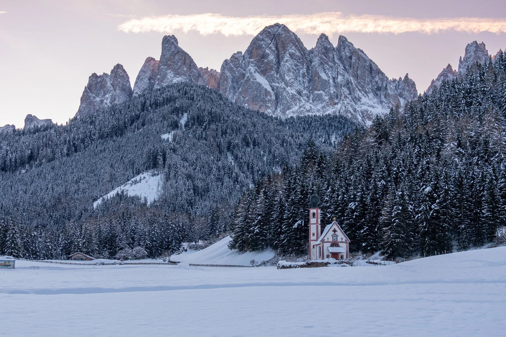 Snow-covered landscape with mountains in the background, a forested hillside, and a small church with a tower in the foreground.