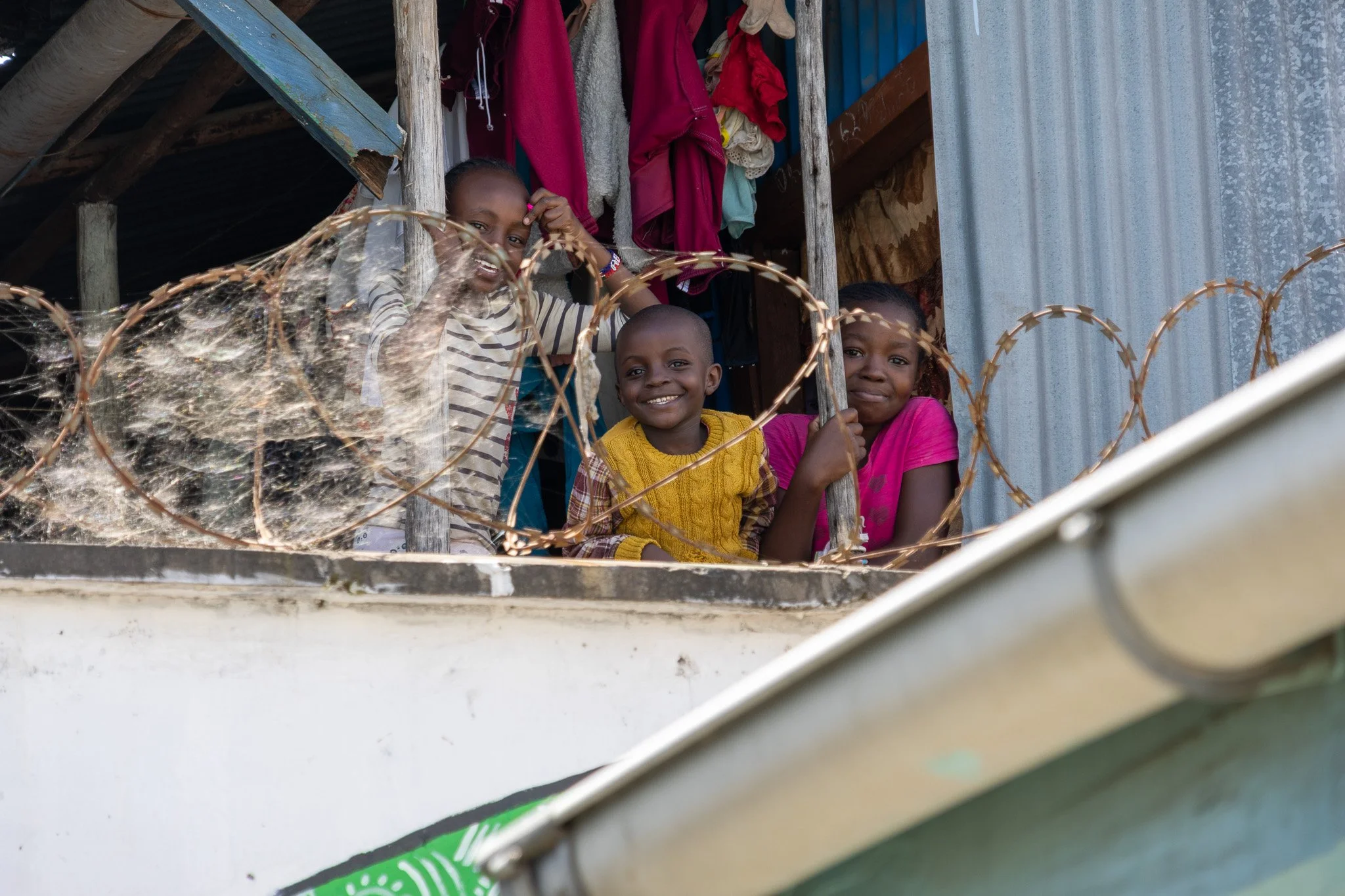 Four children smiling and looking through a barbed wire fence, standing on a balcony of a makeshift structure with hanging clothes behind them.