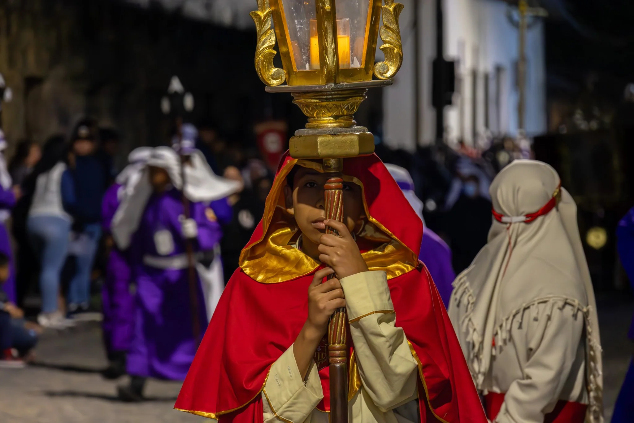 A young person in a red and gold cape holding a lantern during a nighttime parade or procession, with a crowd of people in colorful costumes in the background.