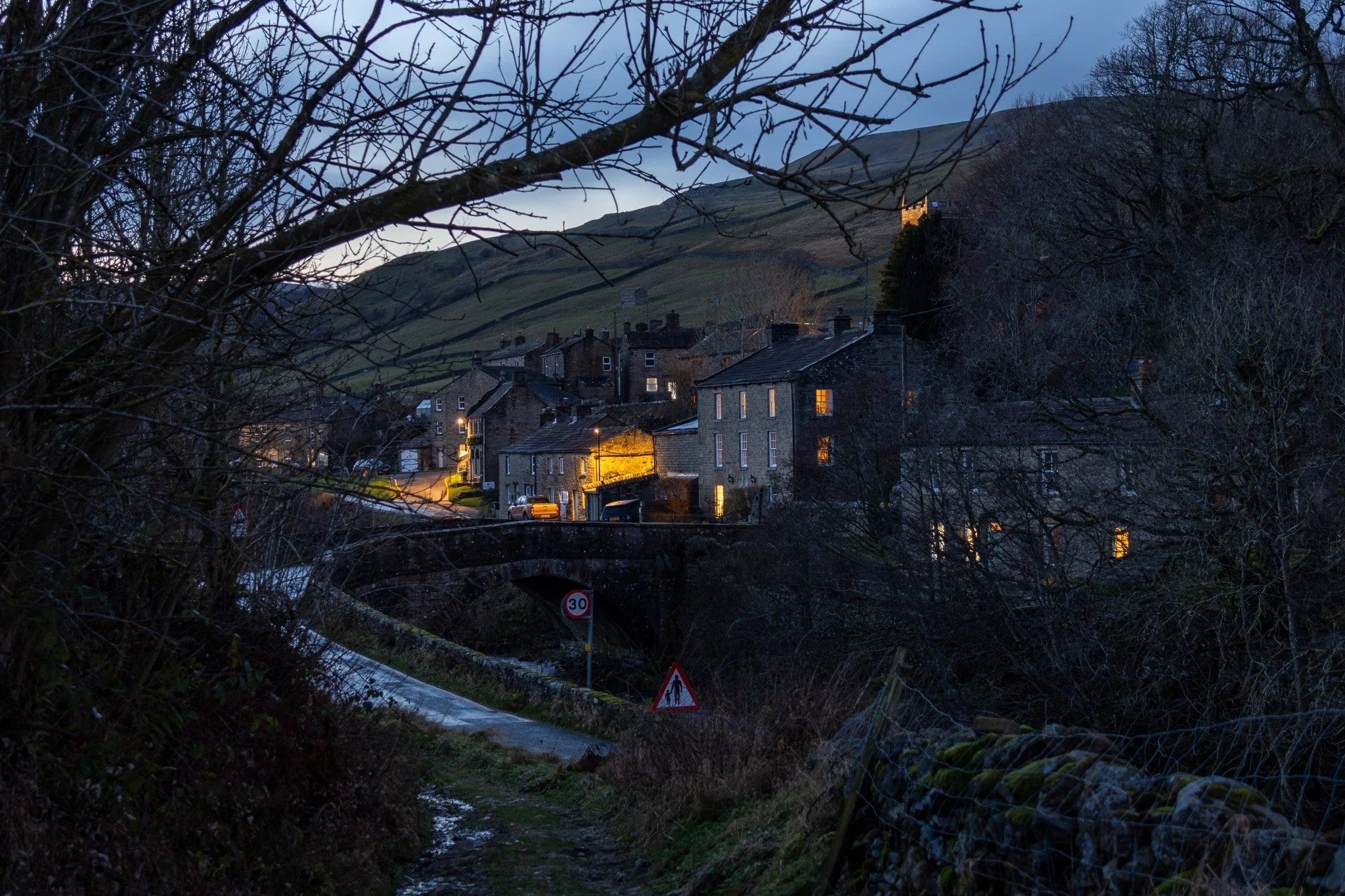 A small village at dusk with houses lit from within, a narrow road, leafless trees, and a hillside in the background.