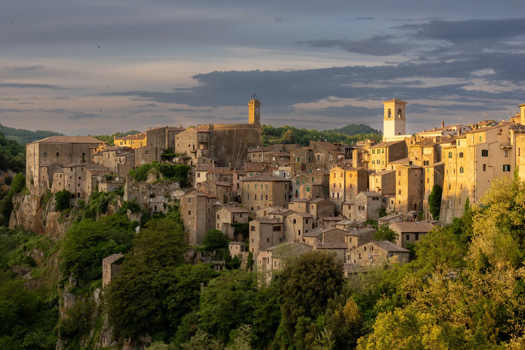 A scenic view of a hillside medieval town with closely packed stone buildings, green trees, and two prominent towers, during sunset with cloudy skies.