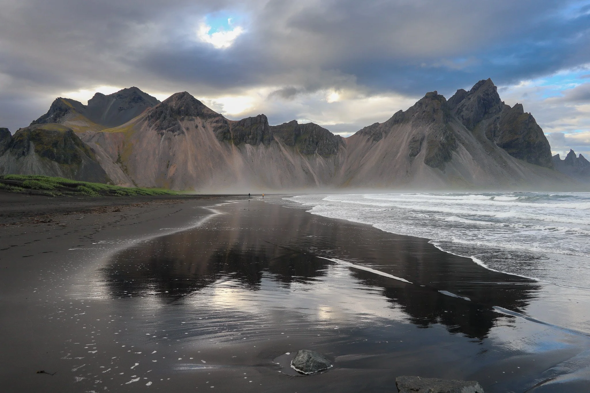 Black sand beach with large rocks, waves, and towering mountains in the background under cloudy sky.