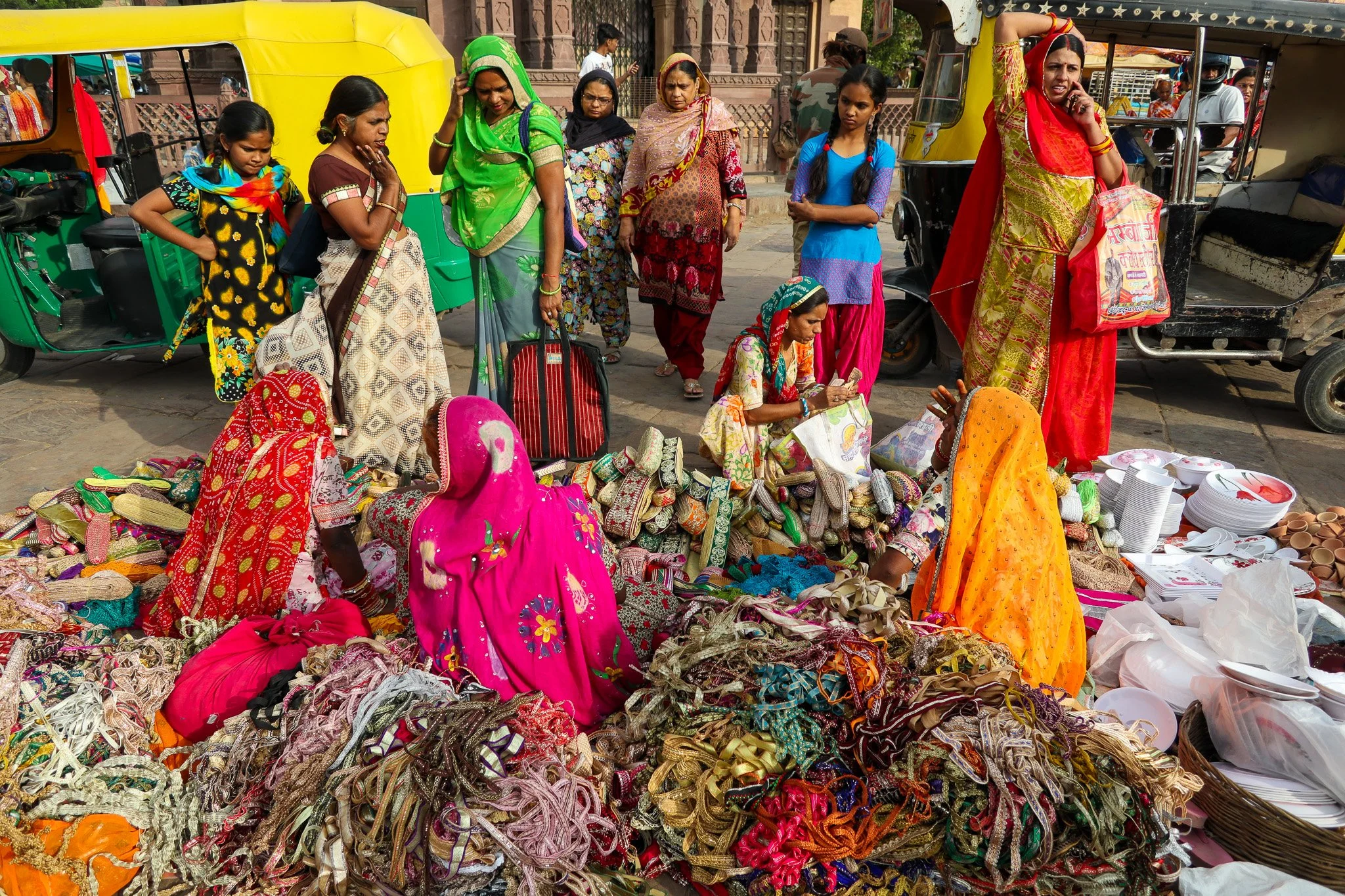 A busy street market scene with women and girls shopping. Women are dressed in colorful traditional Indian attire, with some seated on the ground selling various fabrics, clothing, and accessories, while others stand around, observing and shopping. A