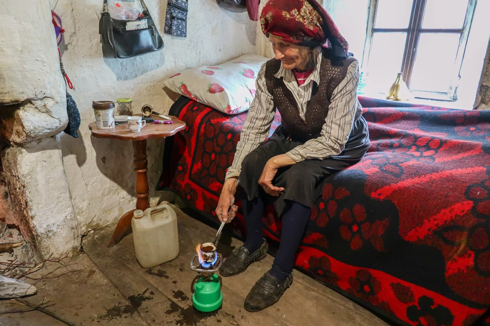 An elderly woman in a headscarf sits on a bed in a rustic room, heating water on a small portable stove, with a table beside her holding a cup, jar, and personal items, while sunlight streams through a window.