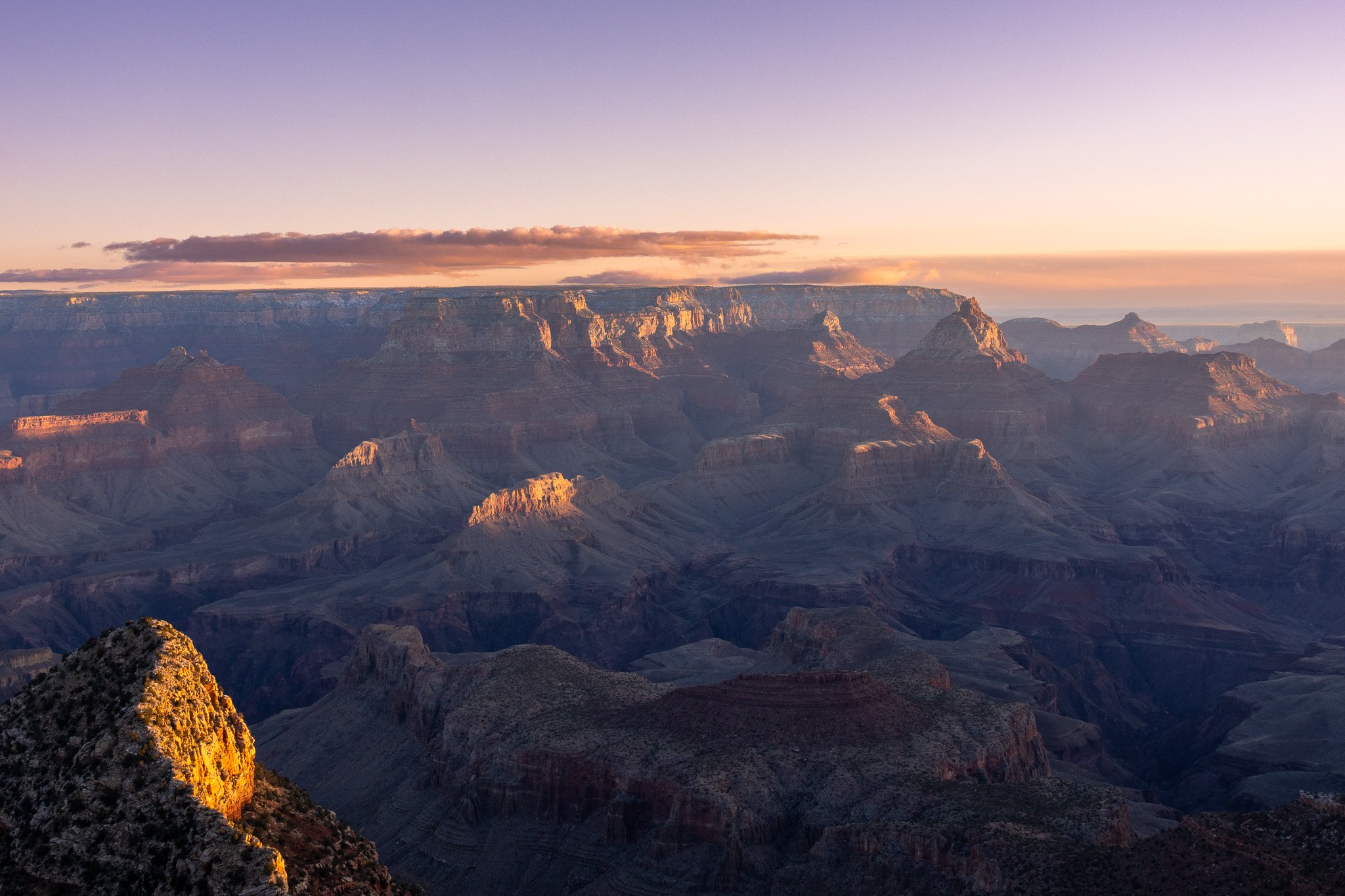 Sunset over the Grand Canyon with soft light illuminating the canyon's layered rock formations and a partly cloudy sky.