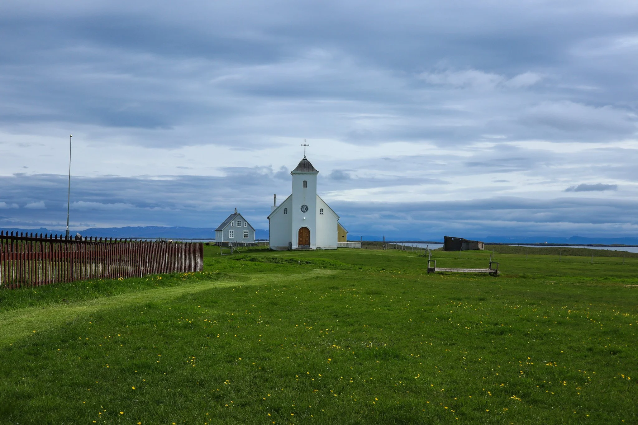 A white church with a steeple and cross, two smaller buildings nearby, set on a green grassy field with yellow flowers, under a cloudy sky.