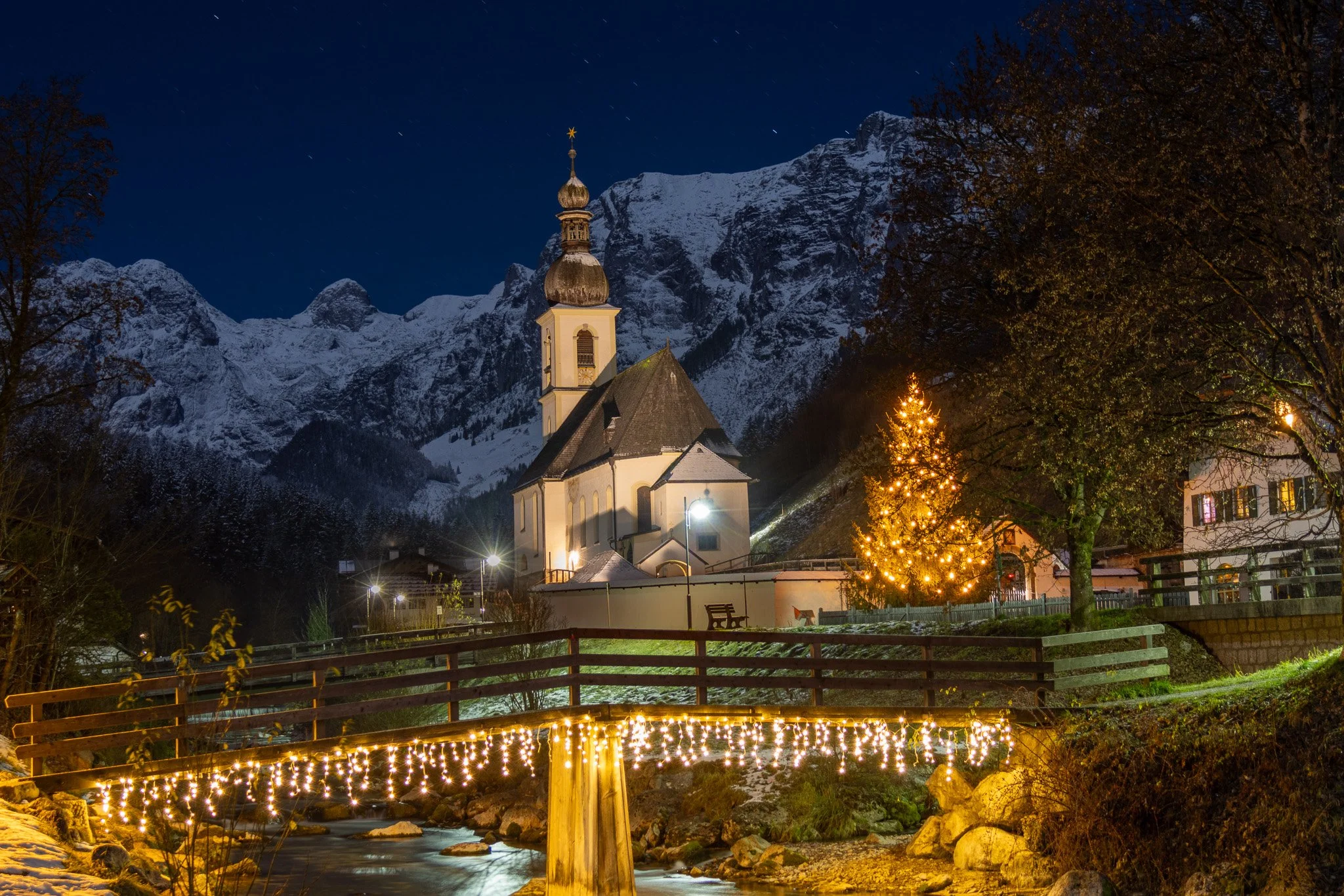 A snowy mountain village at night with a church, decorated Christmas tree with lights, and a small bridge with string lights over a stream.