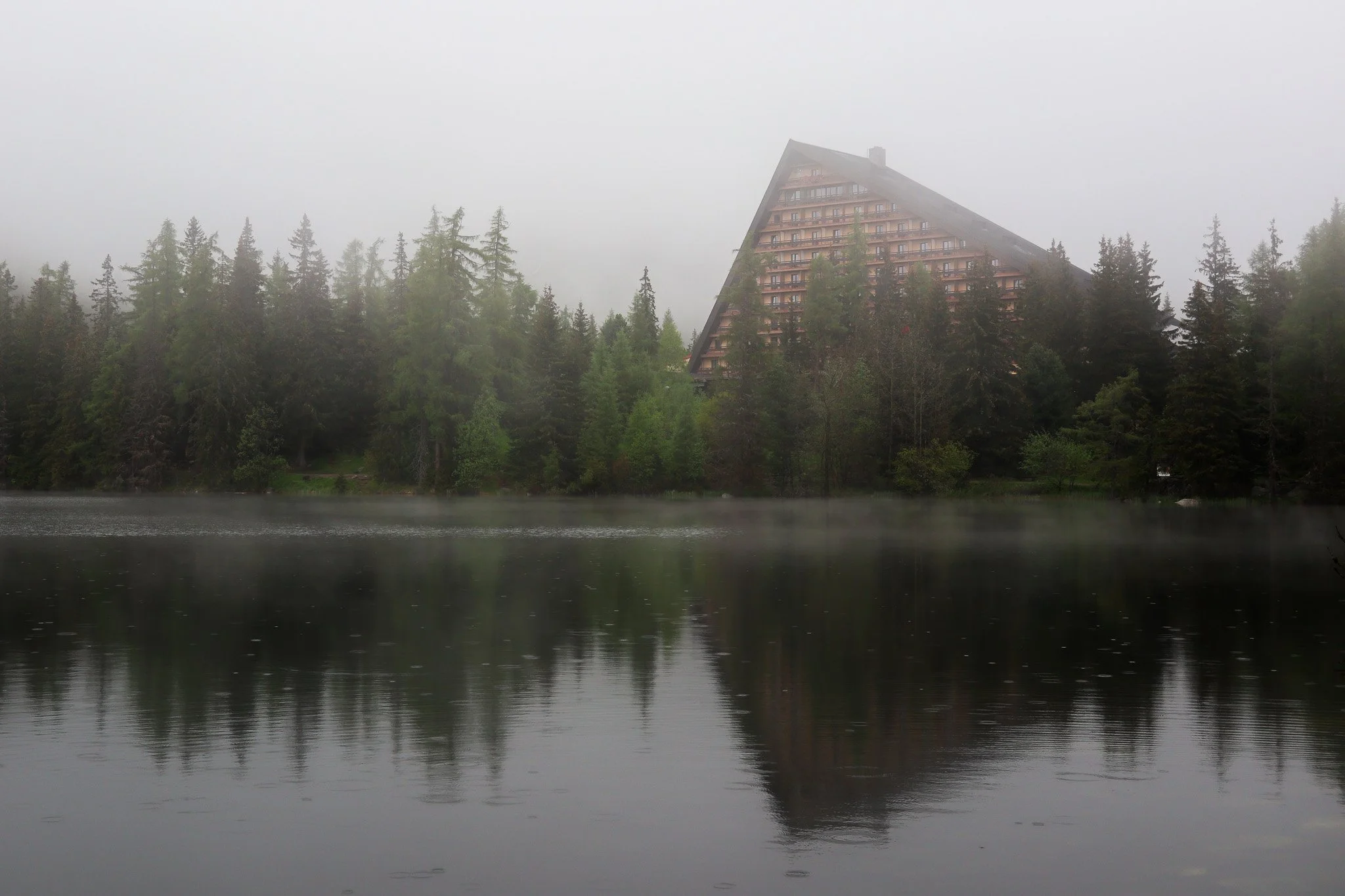 A foggy scene of a lake surrounded by dense green trees with a large triangular-shaped building on the hillside in the background, and its reflection visible on the water.