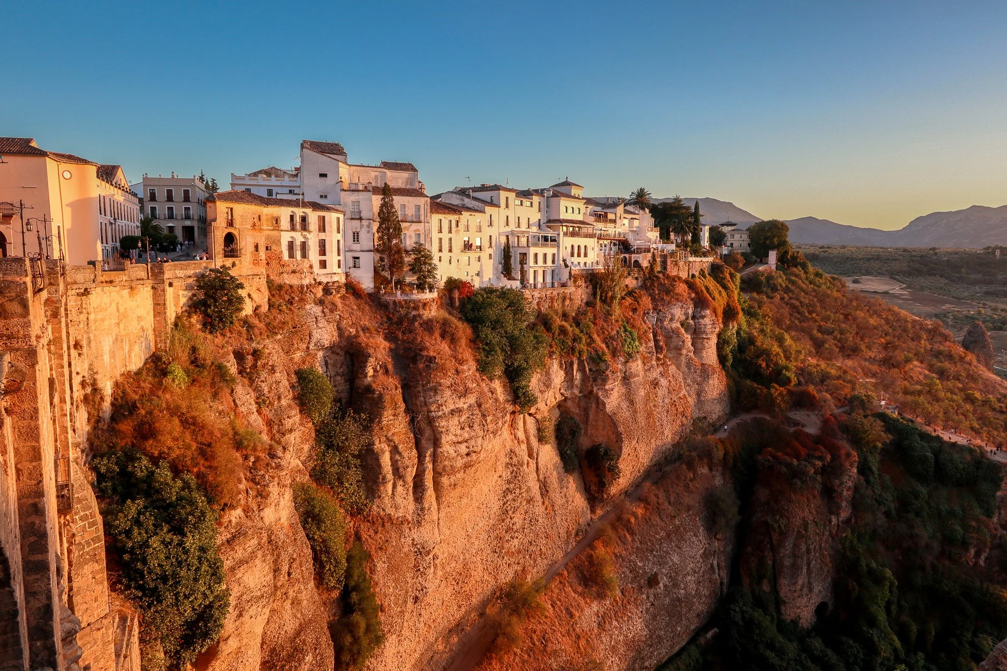 Cliffside town with white buildings overlooking a steep rocky drop at sunset with mountains in the distance.