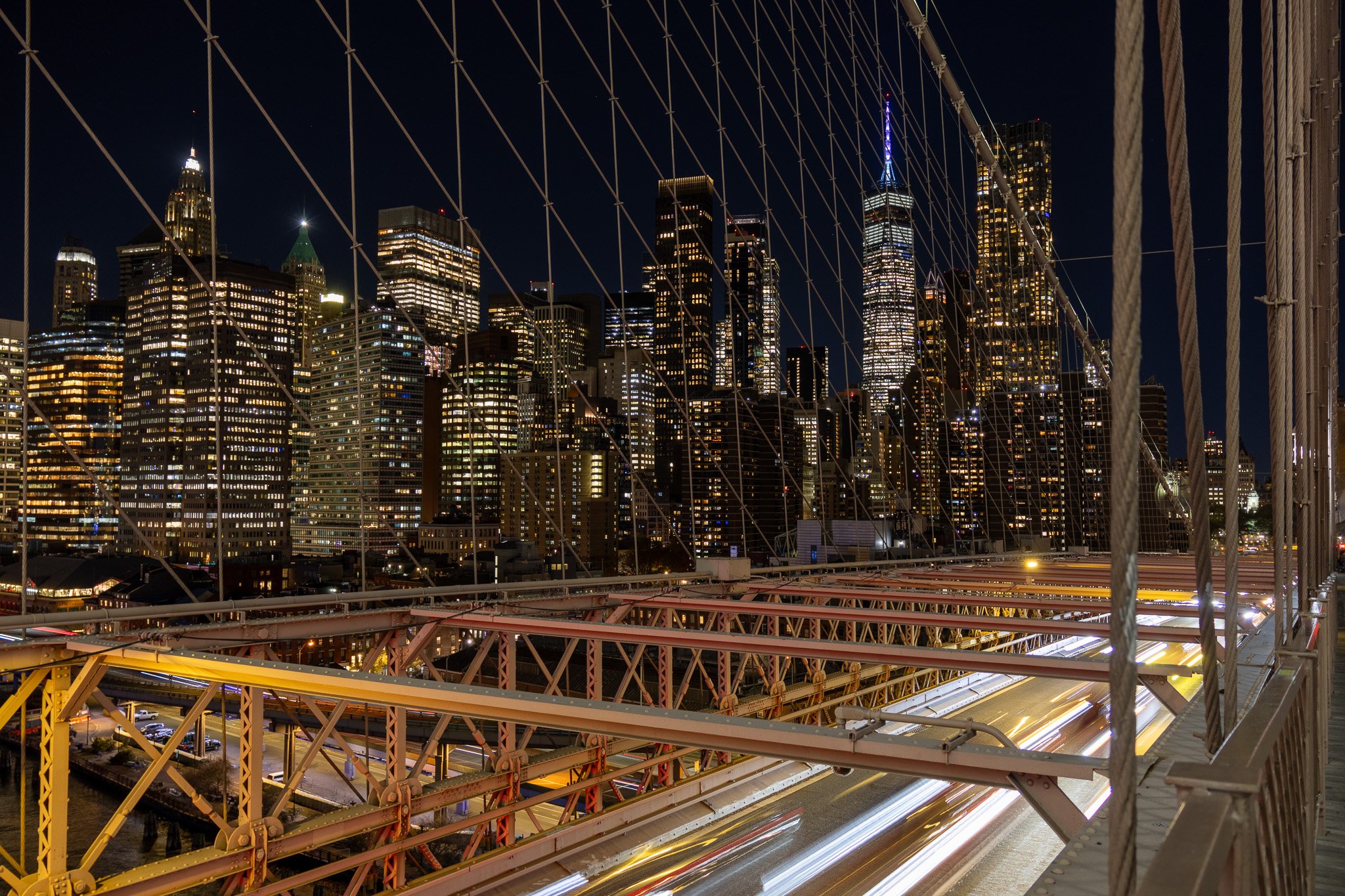 Night view of New York City skyline from Brooklyn Bridge, with light trails on bridge road and illuminated skyscrapers including One World Trade Center.